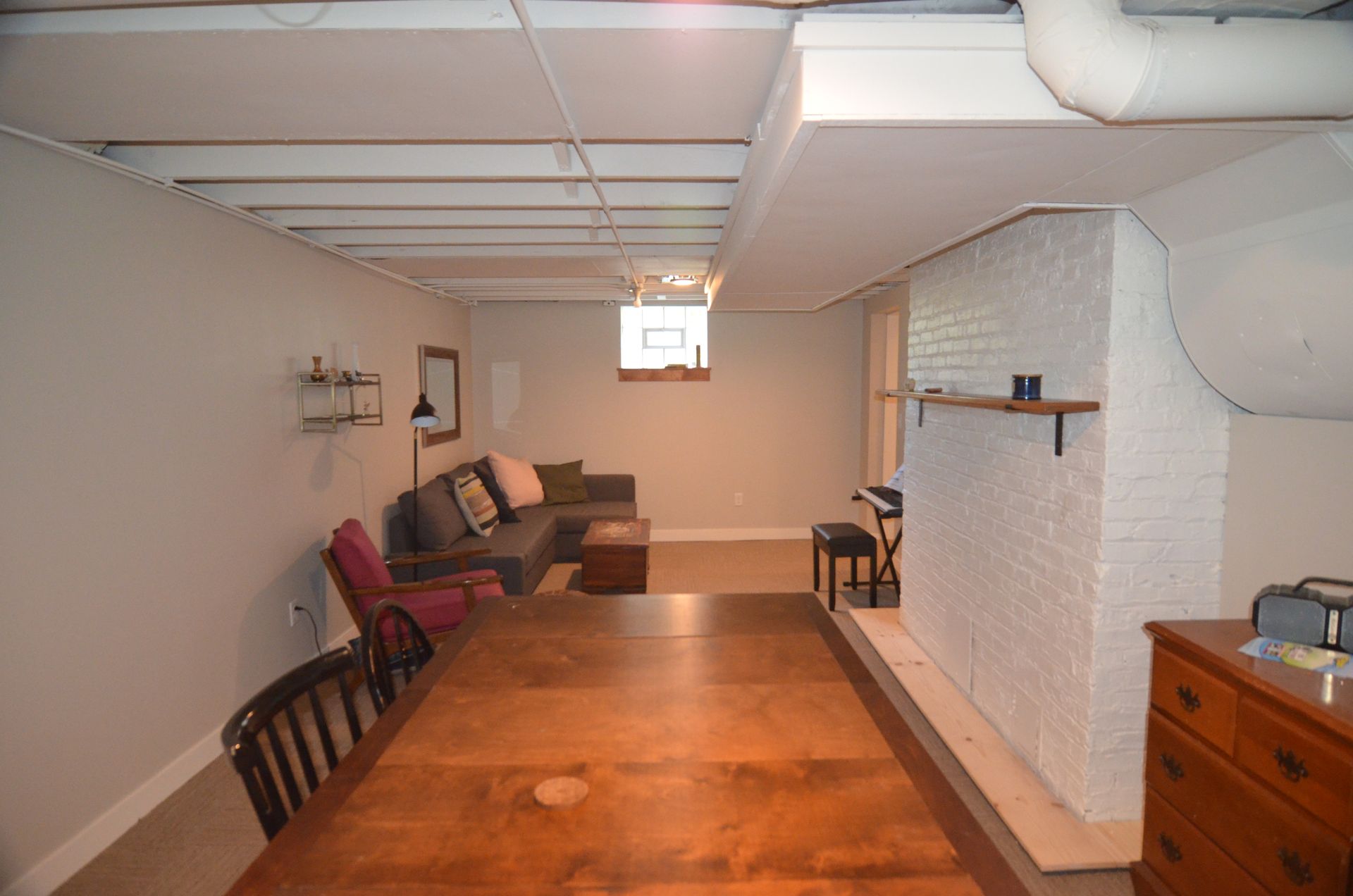 Basement room with long wooden table, sofa, and painted brick wall; neutral colors.