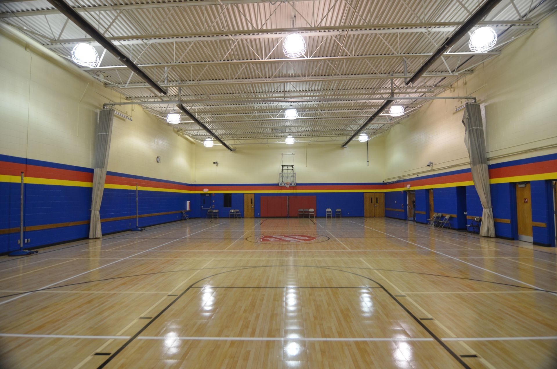 Empty gymnasium with hardwood floor, basketball hoop, and blue and yellow walls.