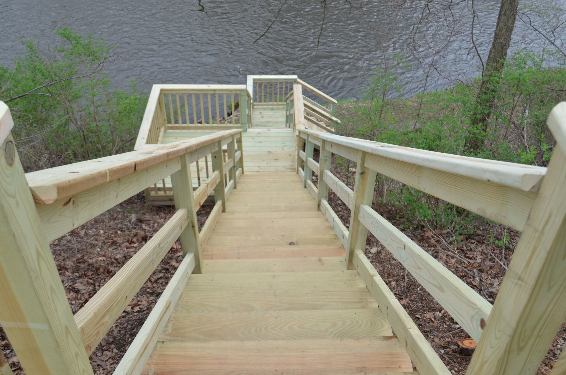 Wooden staircase leading down to a river. Handrails on both sides, with a small landing at the top.
