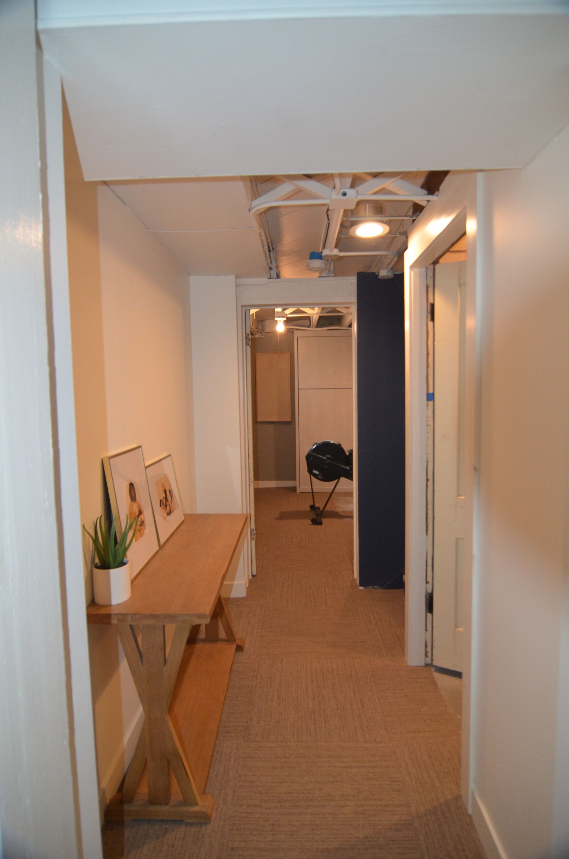Narrow hallway with light-colored walls and carpet; wooden table with framed art on the left; doors on the right.