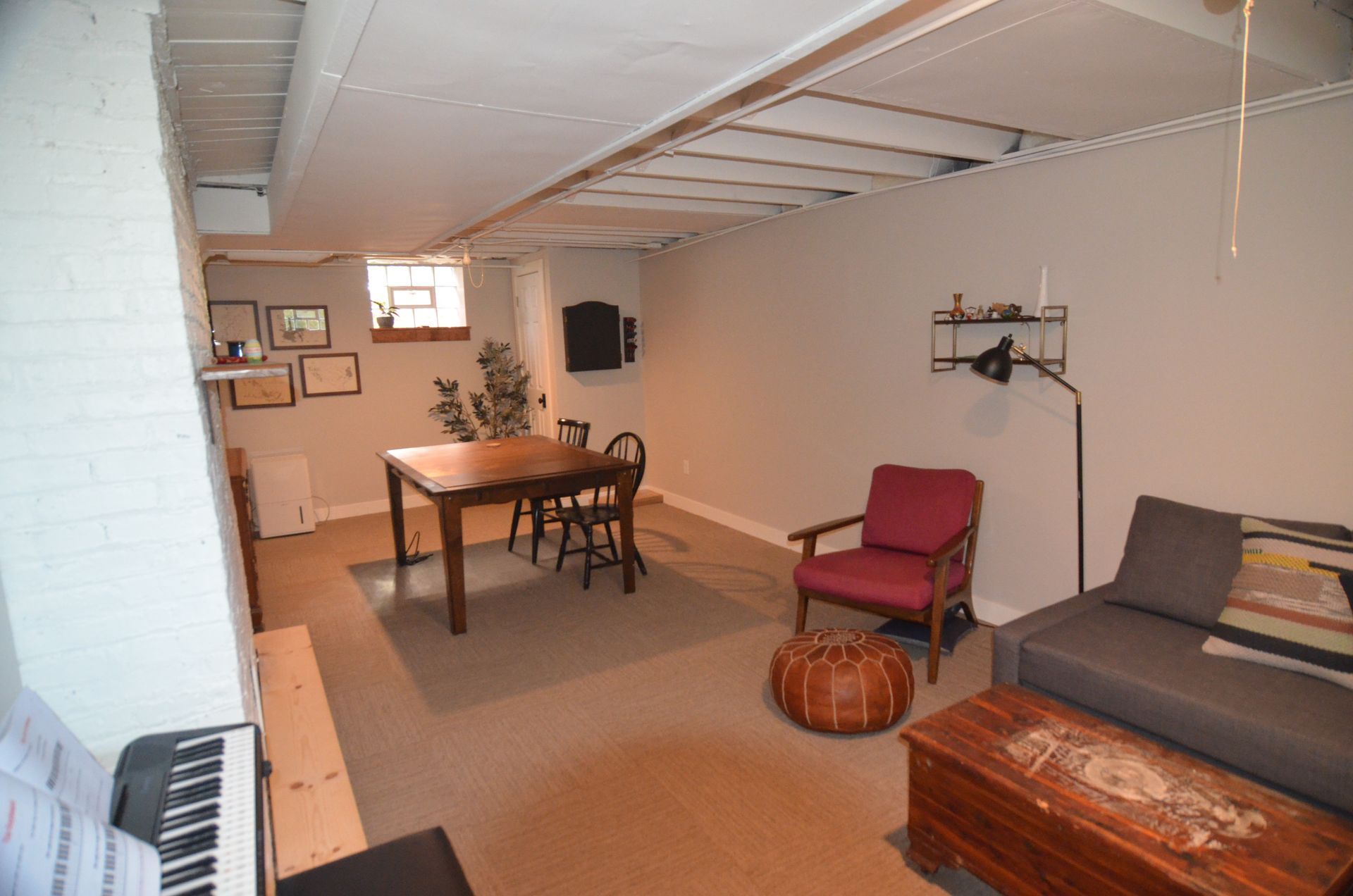 Basement room with table, chairs, sofa, chair, ottoman, and keyboard. Beige walls and carpet.