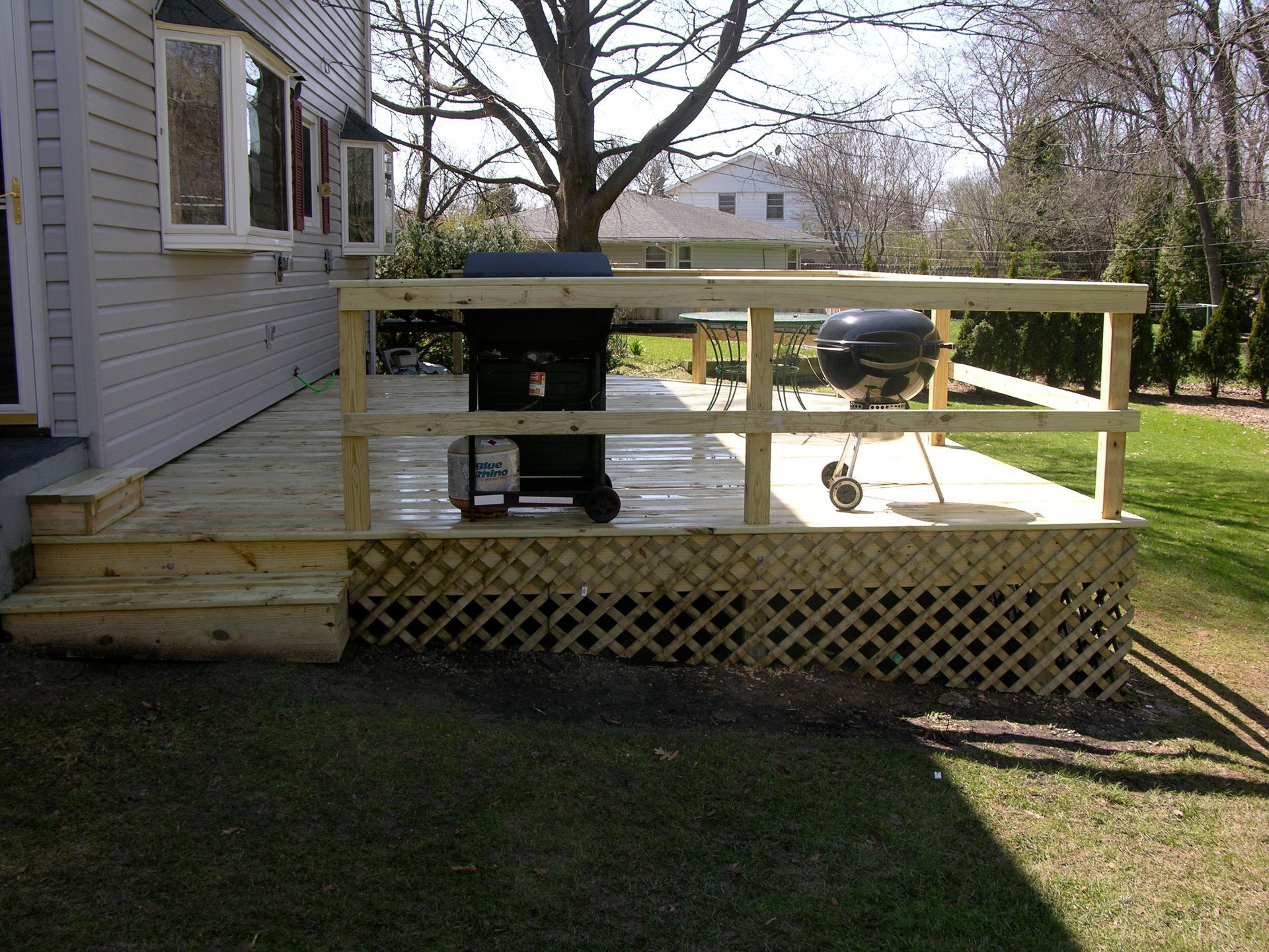 Wooden deck with grill and railing, next to a house with a bay window and steps on a grassy lawn.
