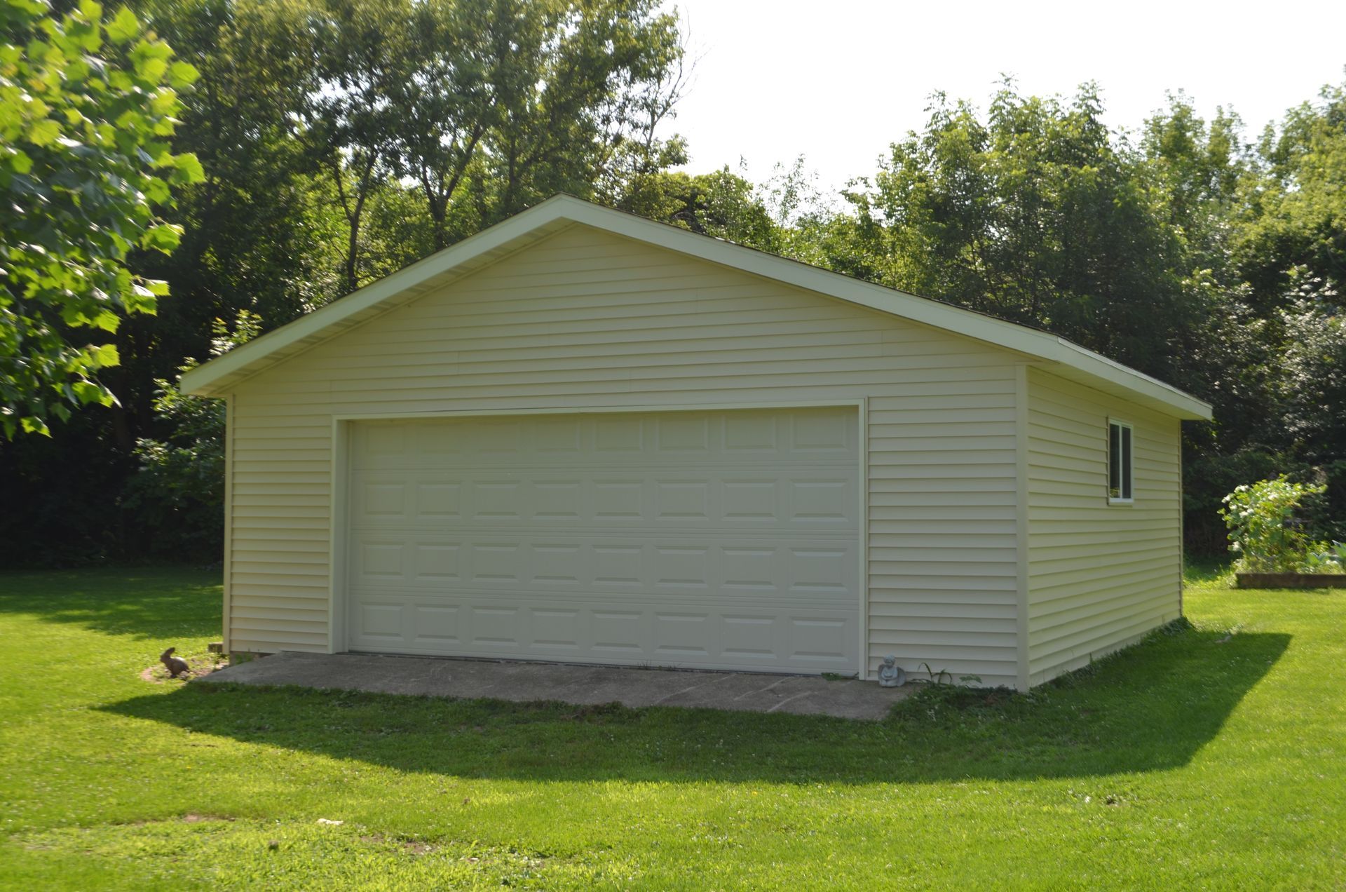 Cream-colored detached garage with a closed garage door, on a green lawn, surrounded by trees.