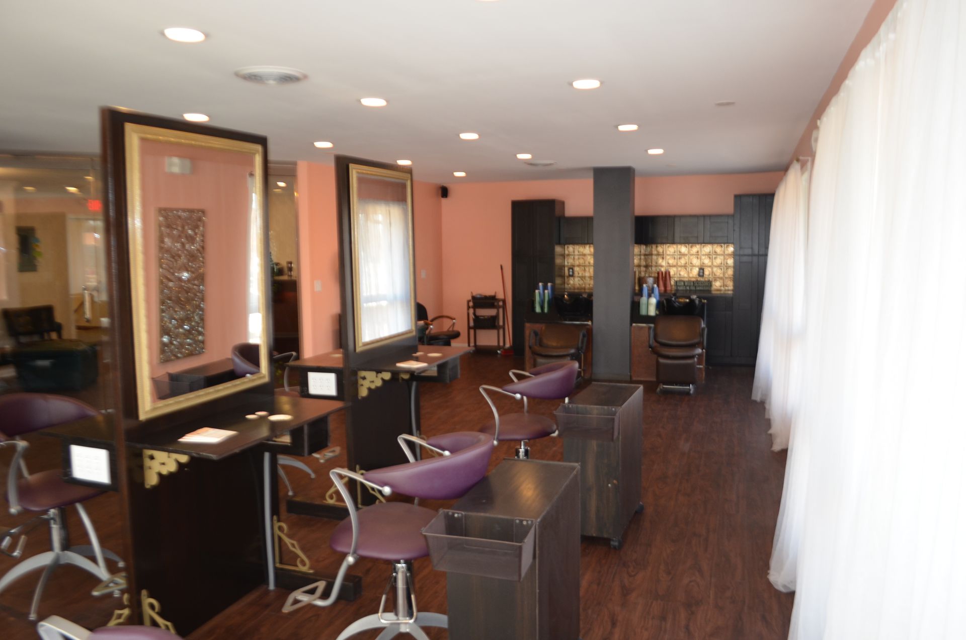 Hair salon interior with styling stations, mirrors, and chairs. Pink walls, dark wood floors.