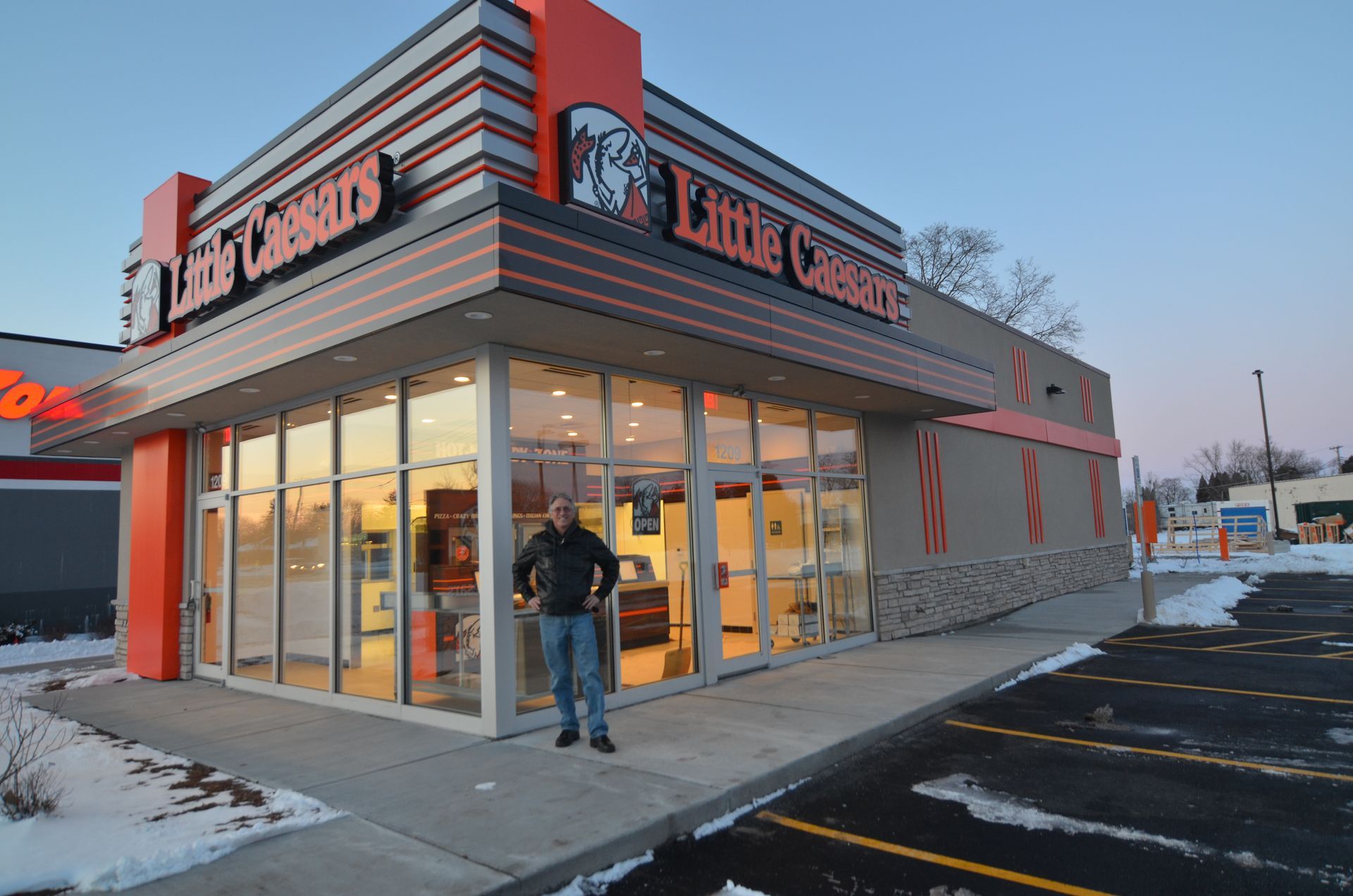 Man stands outside a Little Caesars restaurant. Building is orange and gray with large windows. Snow on the ground.