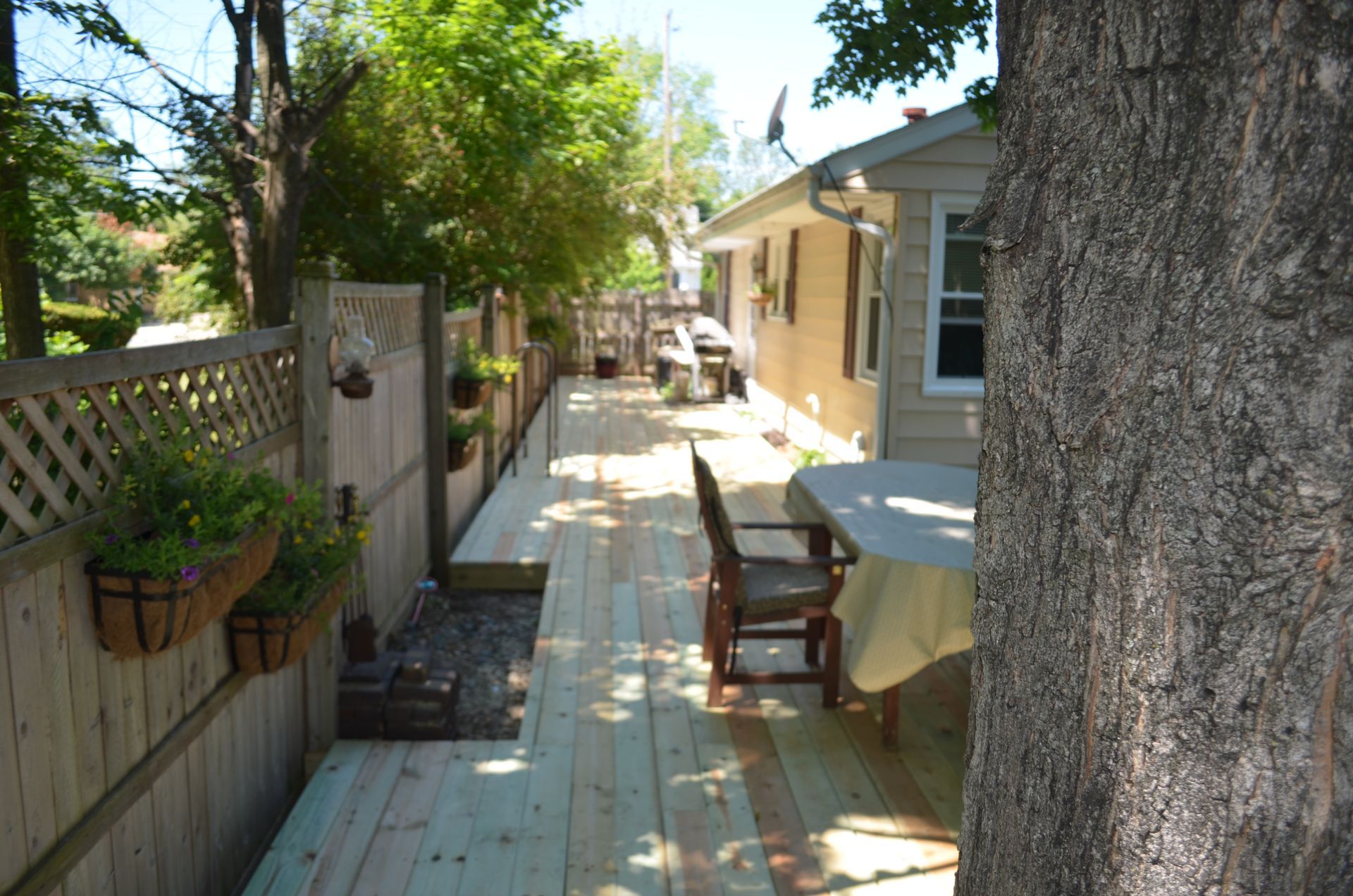 Wooden deck with a fence, hanging flower baskets, and a table beside a house.