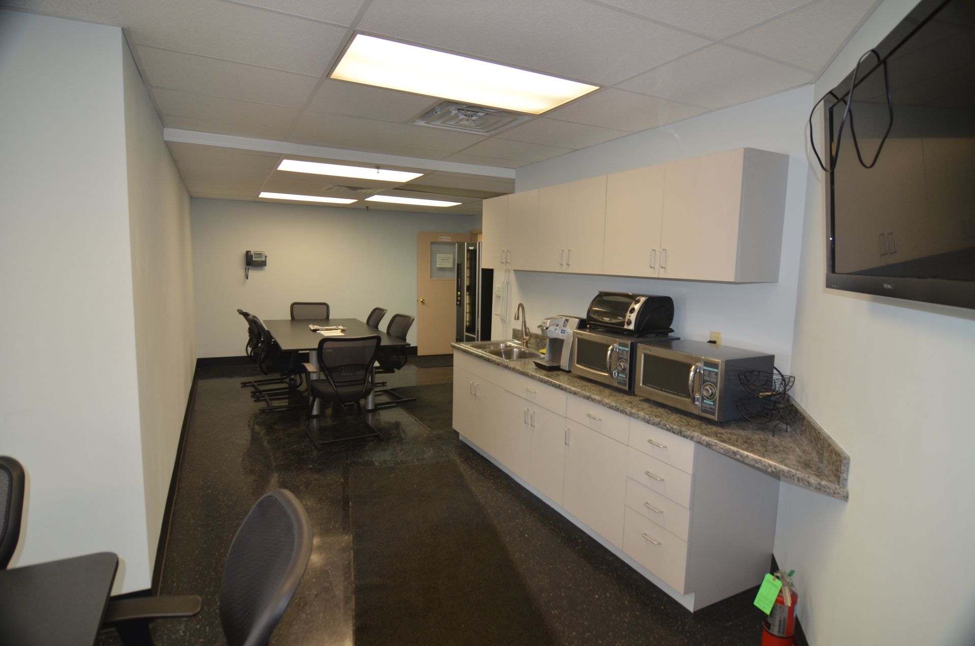 Office space with conference table, kitchenette, and TV. White cabinets and walls. Black floor.