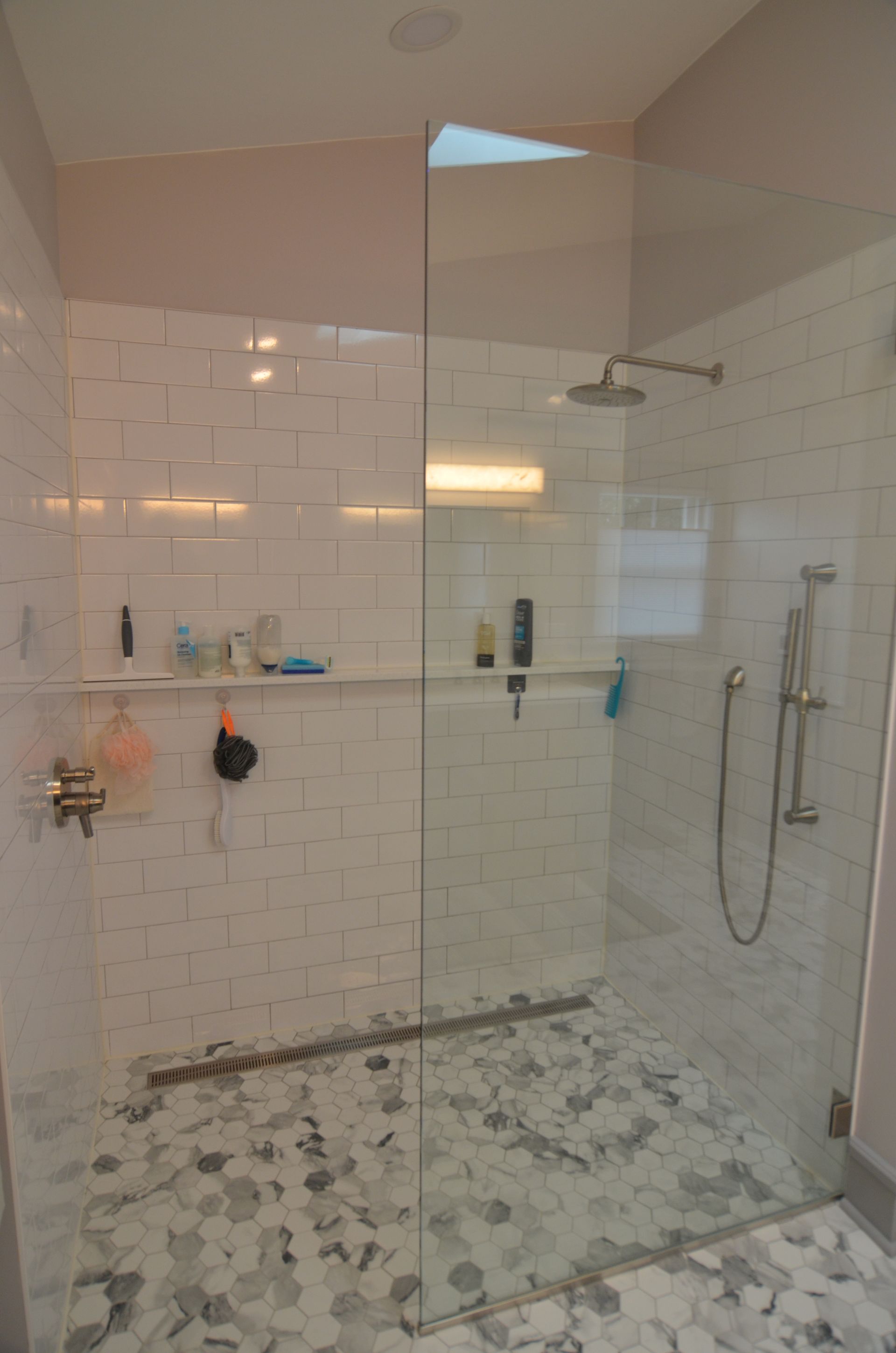 Modern white-tiled shower with glass door and hexagon floor tiles. Contains shampoo bottles and shower heads.