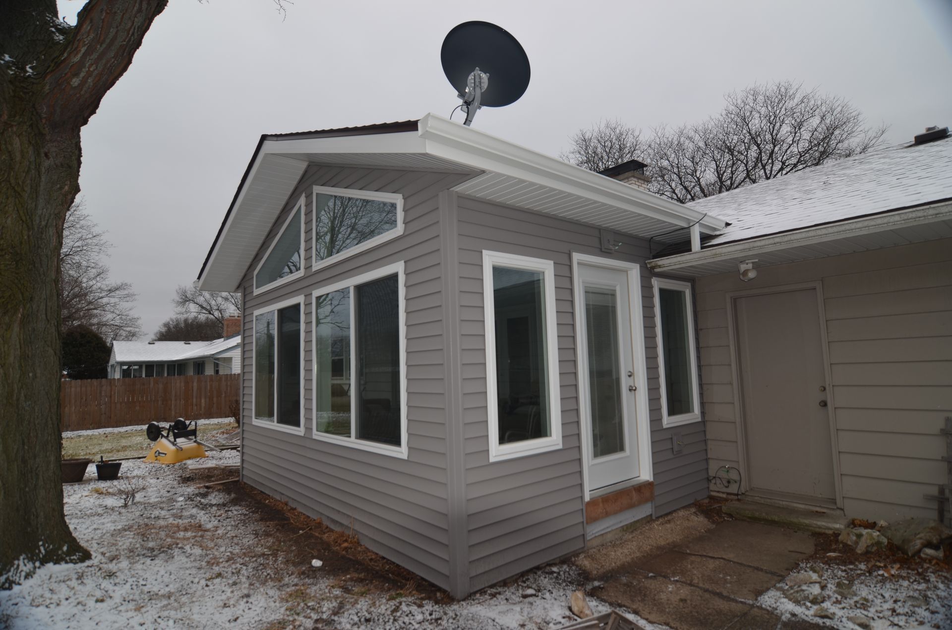 A sunroom addition with gray siding and white-framed windows and a door, attached to a house with a satellite dish on its roof.