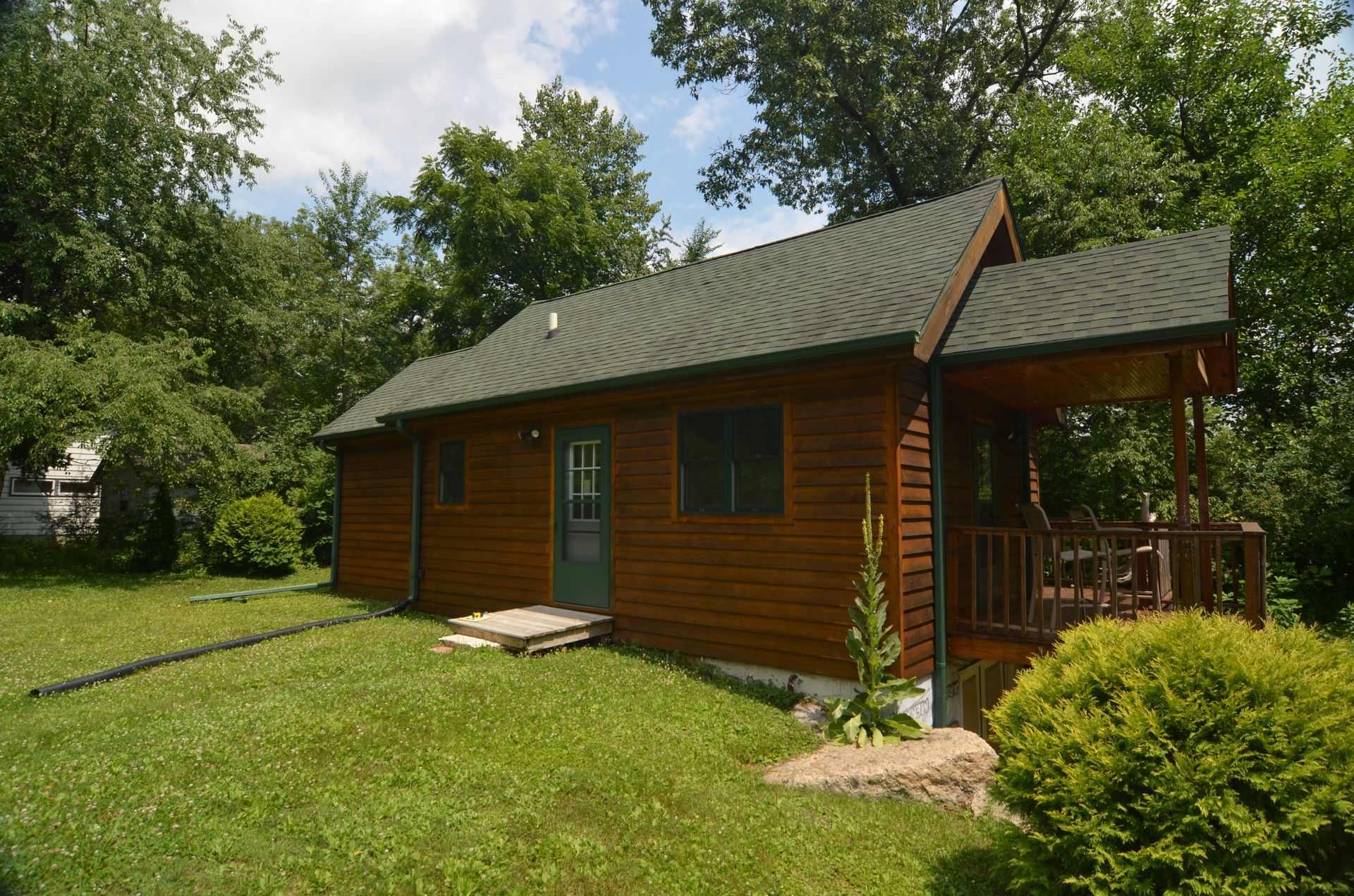 Brown cabin with green roof and small porch, surrounded by trees and grass under a blue sky.