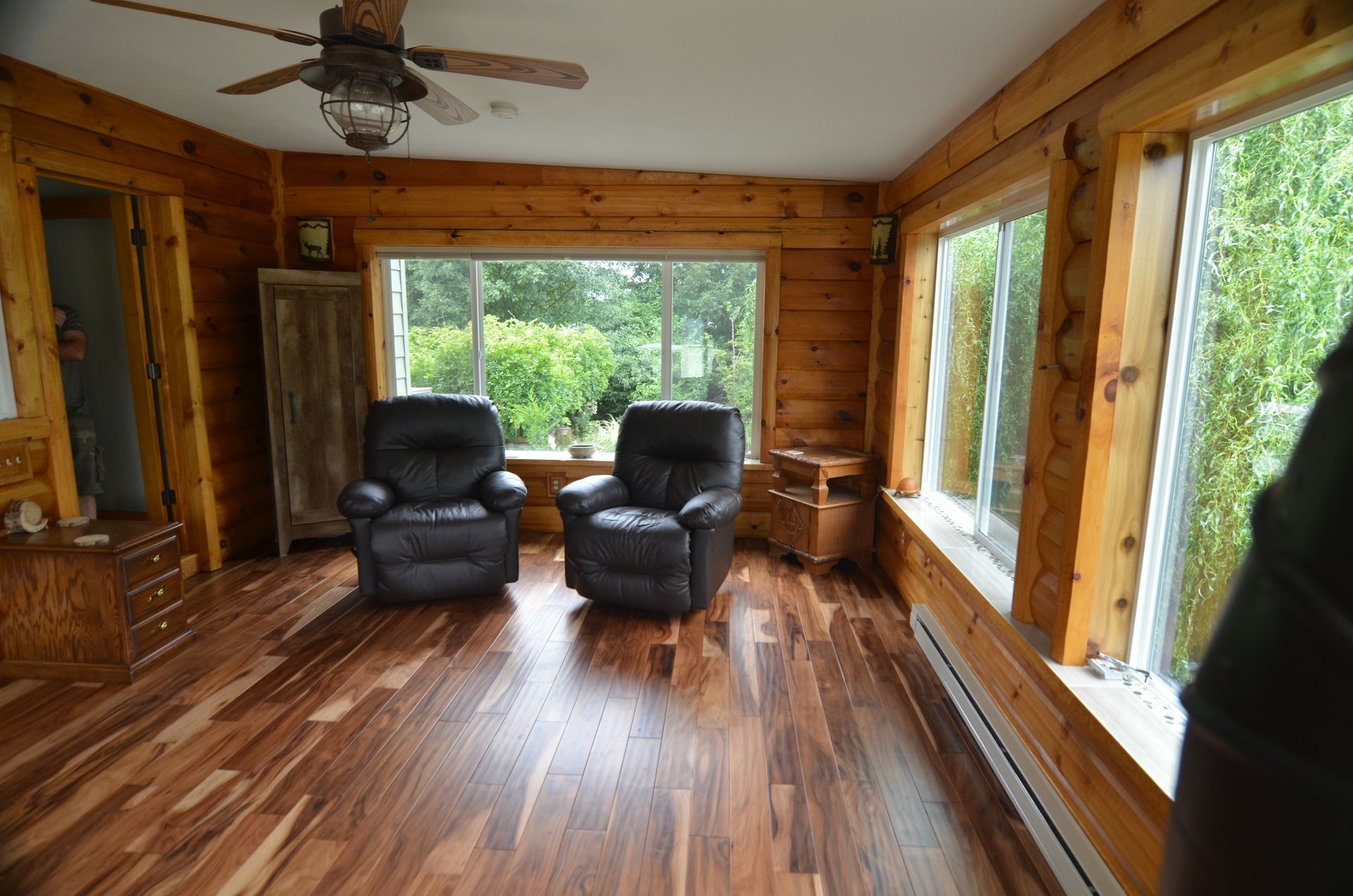 Sunroom with wood paneling, hardwood floor, two black recliners, and large windows.