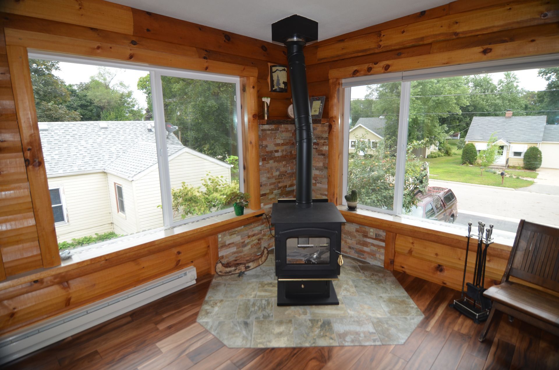 Cozy room with wood stove and corner windows overlooking neighborhood. Wood paneling, black stove, gray tile floor.