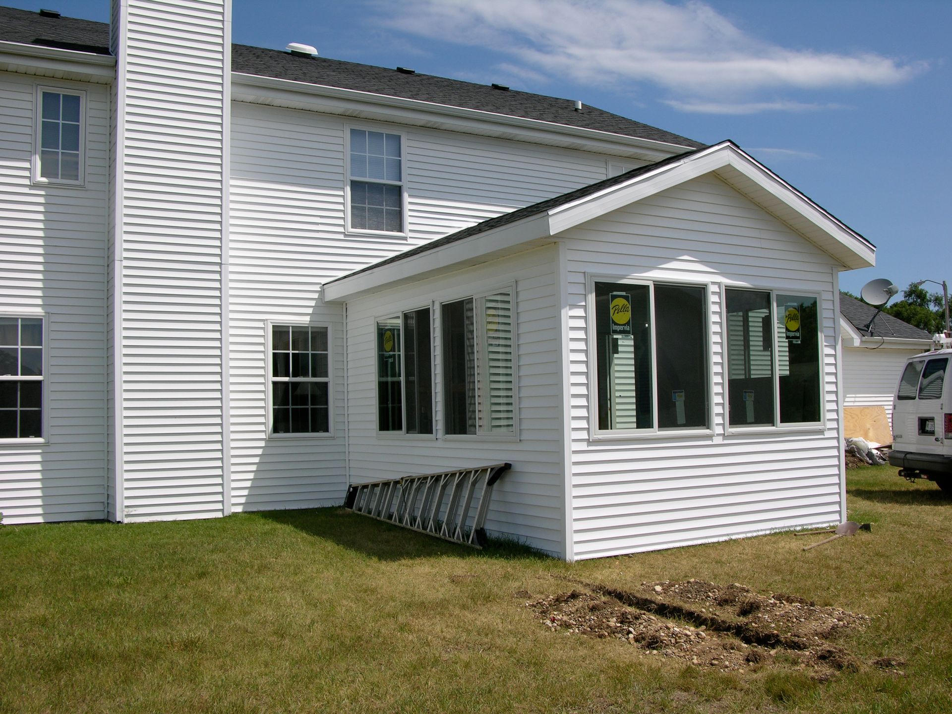 White house with attached sunroom, multiple windows, green grass, ladder leaning against sunroom.
