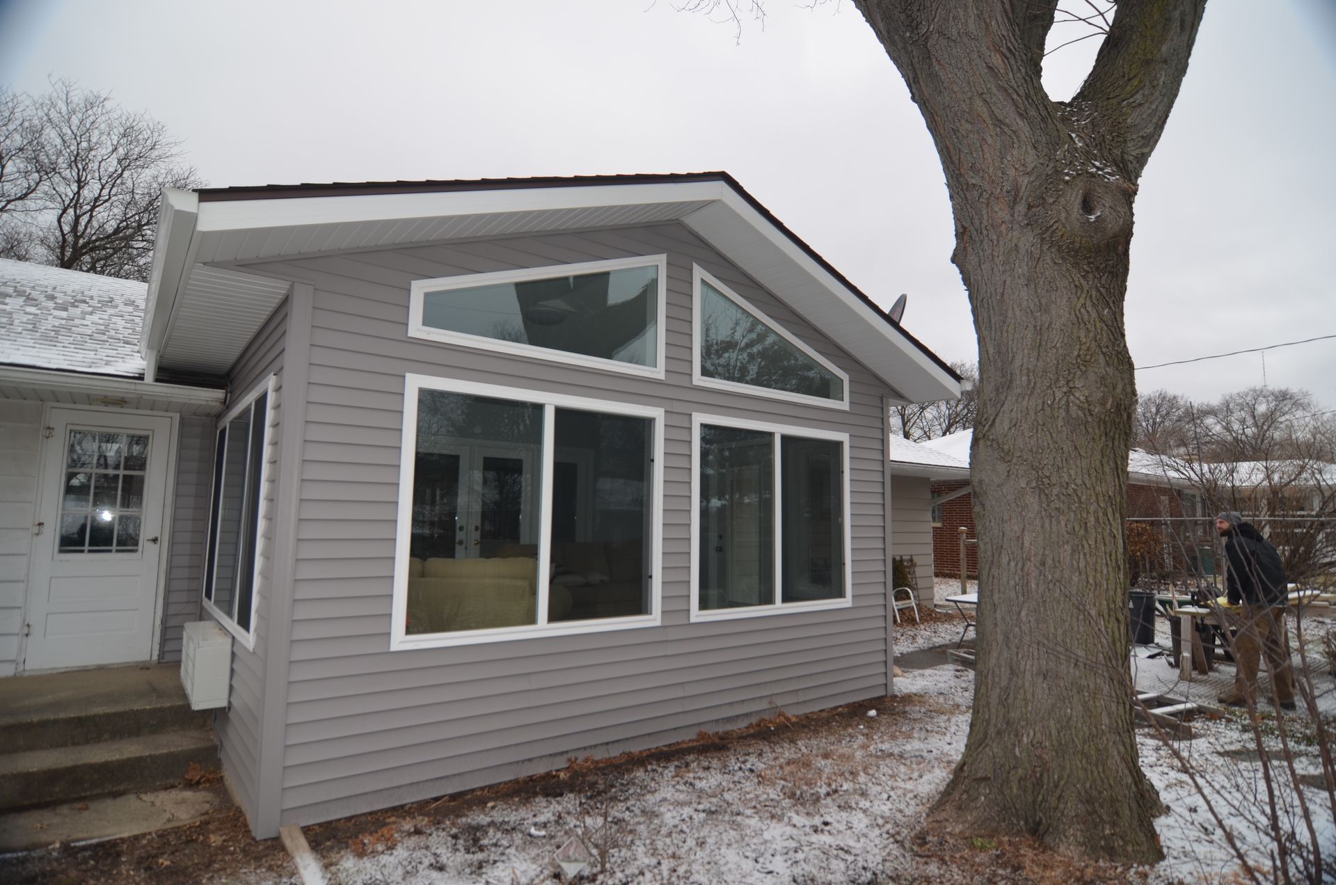 Gray-sided sunroom addition with large windows, attached to a house; snow on ground, cloudy day.