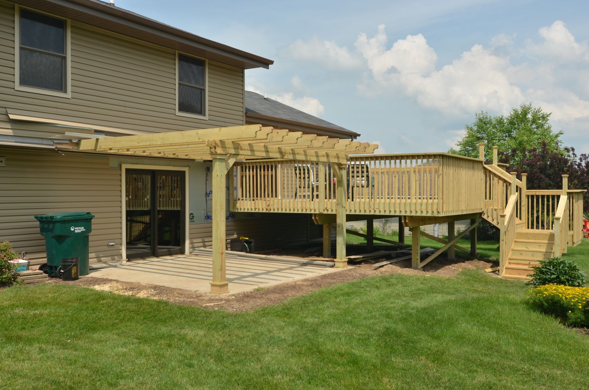 Wooden deck with pergola, stairs, and patio next to a two-story house, set in a grassy yard under a cloudy sky.