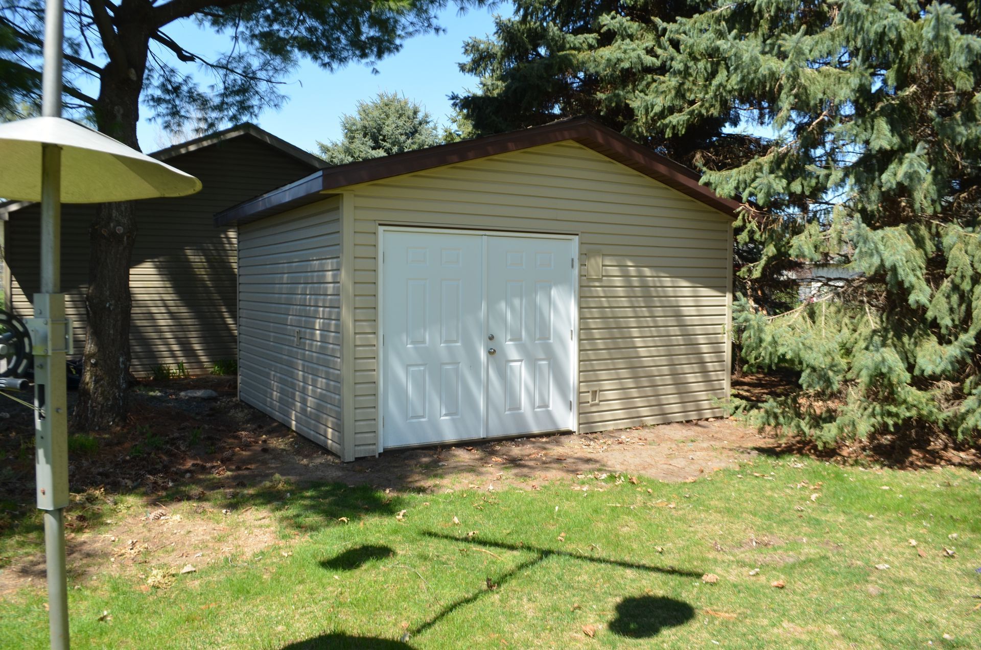Tan shed with white doors in a grassy yard, surrounded by trees under a blue sky.