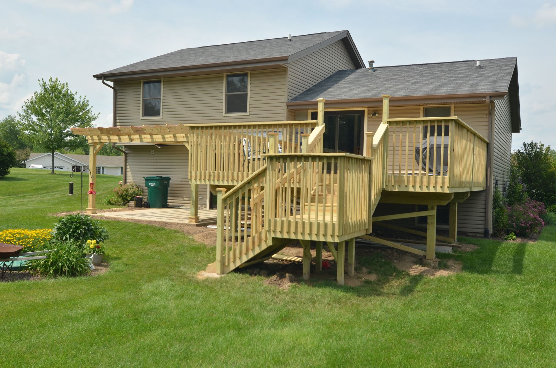 Two-story home with wooden deck and pergola. Green lawn and a small garden. Sunny, daytime.