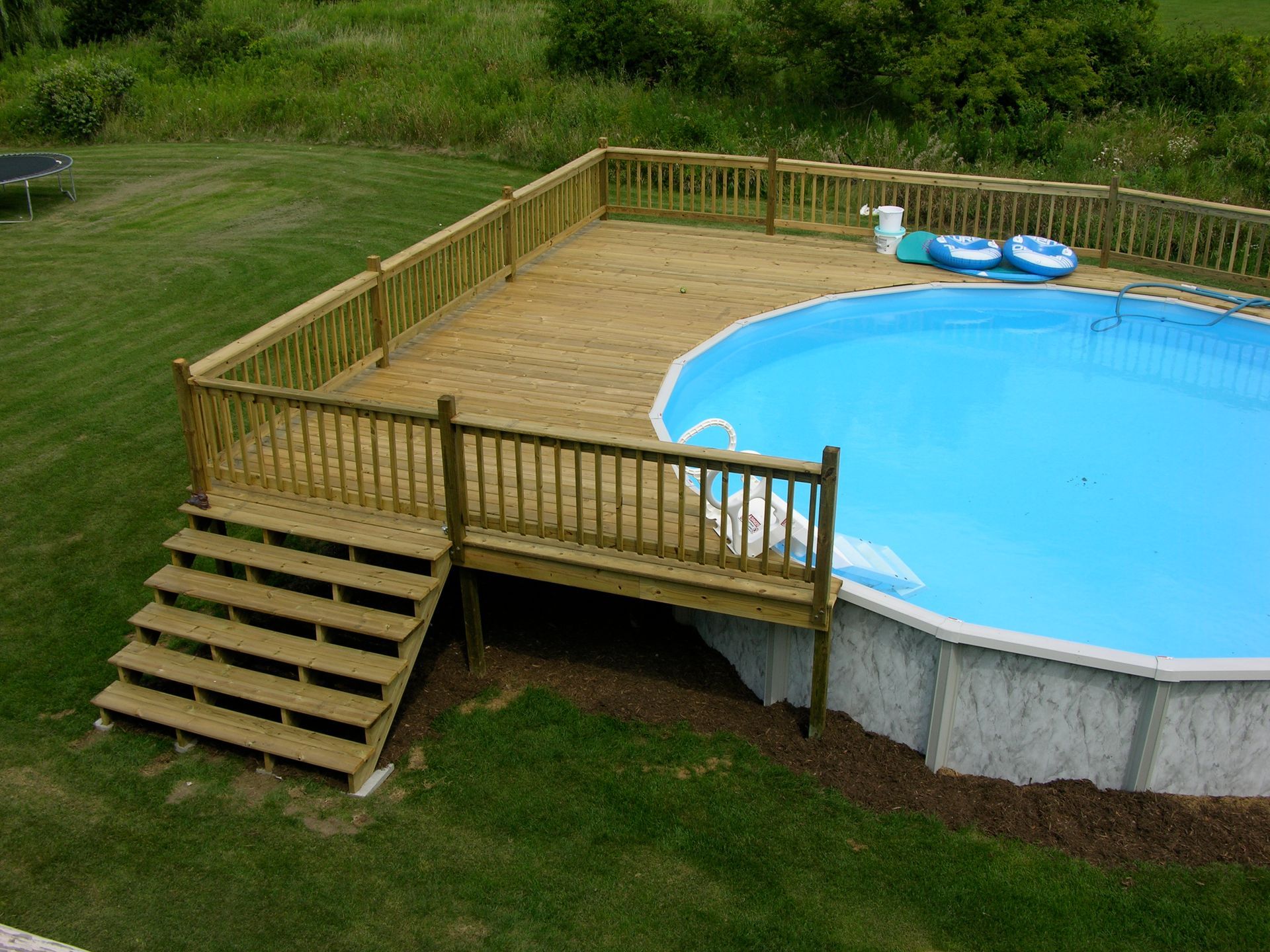 Wooden deck surrounds an above-ground pool. Stairs lead to the deck, which has a railing.