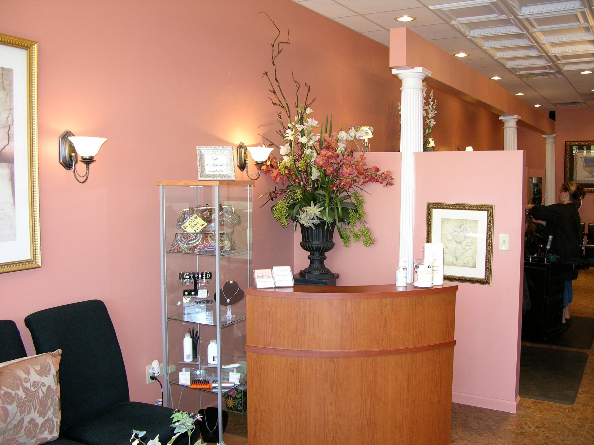 Reception area of a hair salon; pink walls, wooden desk, floral arrangement, glass display case, waiting chairs.