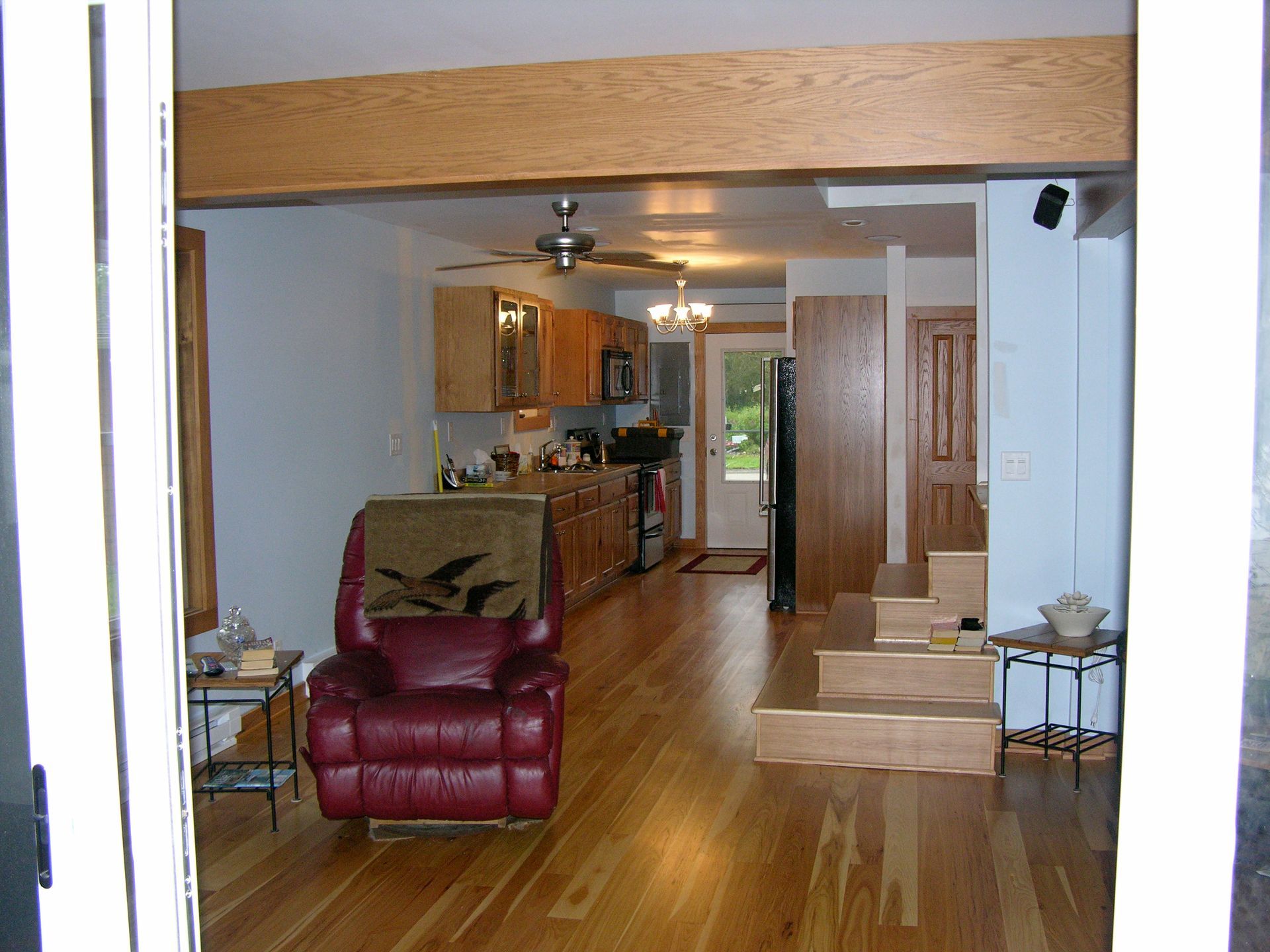Interior view of a home with hardwood floors, kitchen, recliner, and stairs leading to a door.