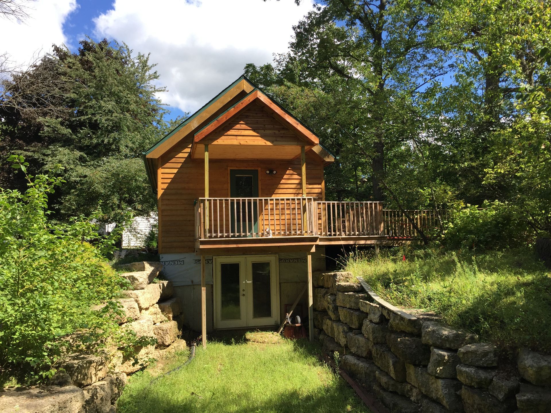 Small wooden cabin with a deck, built into a hillside with a stone retaining wall.