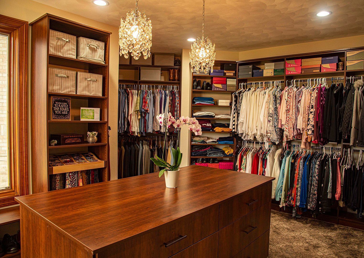Walk-in closet with island, shelves, and hanging clothes. Two chandeliers illuminate the space.