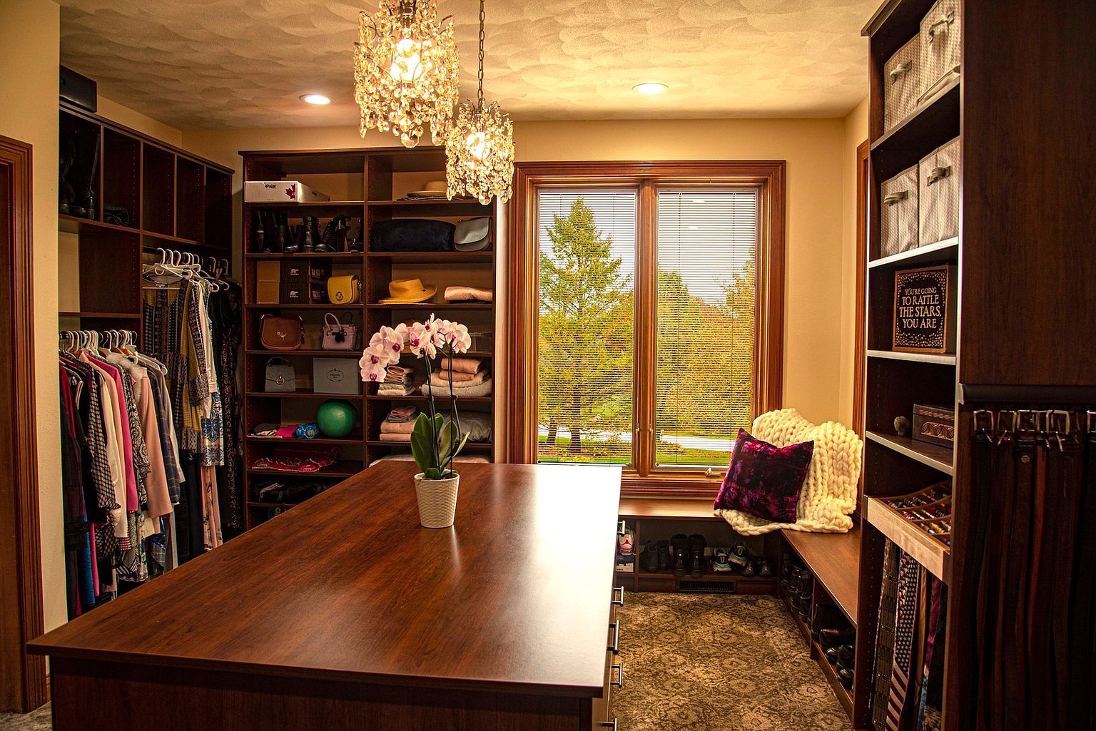 Walk-in closet with dark wood shelving, island, window seat, and hanging clothes.