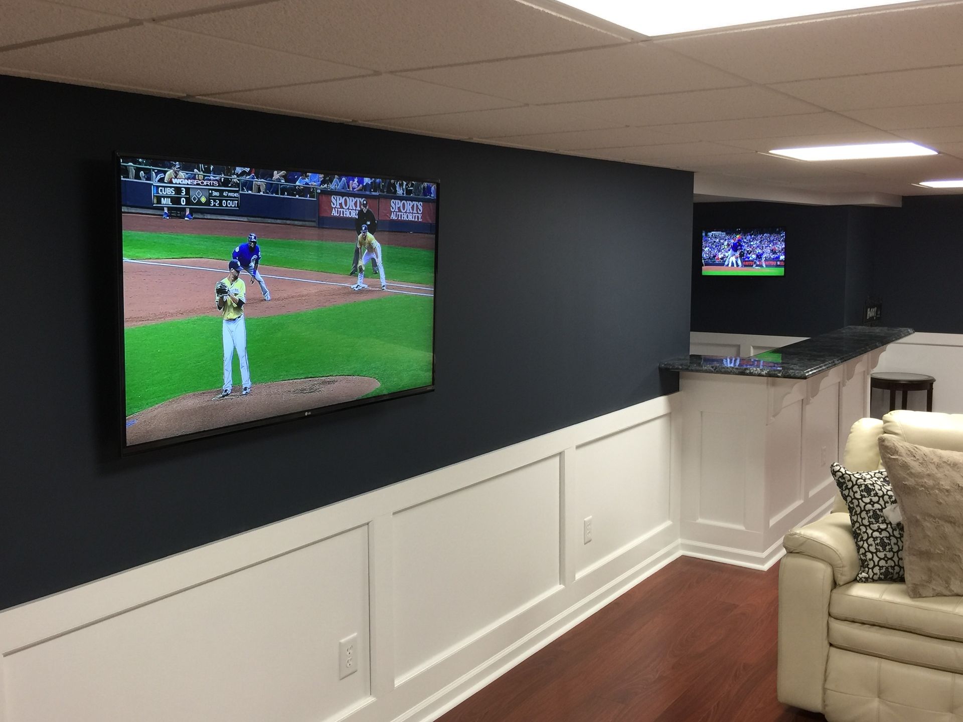 Basement entertainment area with two TVs showing baseball game. Dark blue wall, white trim, wood floor.