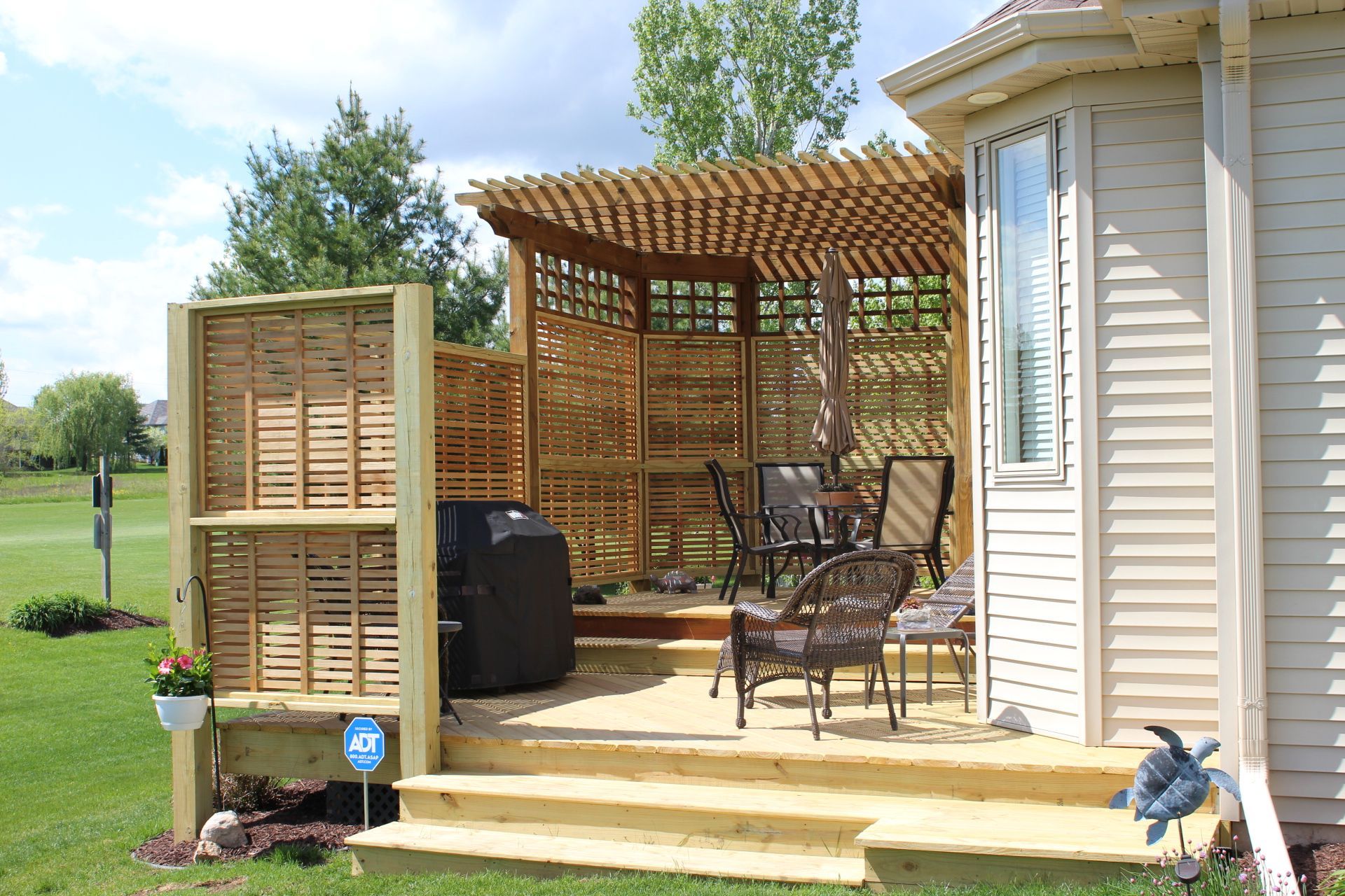 Wooden deck with pergola, privacy screens, and outdoor seating next to a beige house, with greenery and lawn.