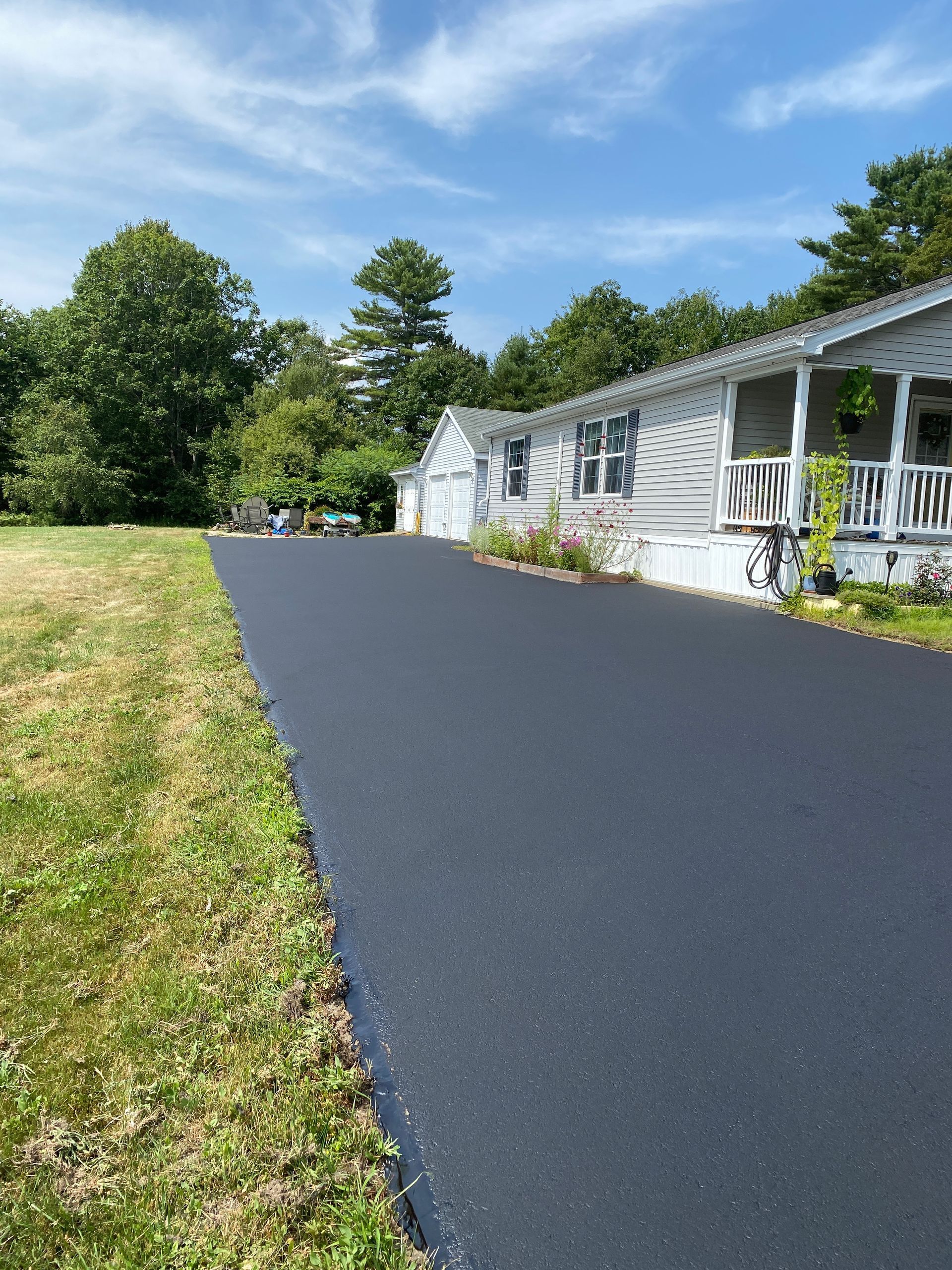 A freshly paved black asphalt driveway leading up to a light gray house with a white porch under a blue sky.