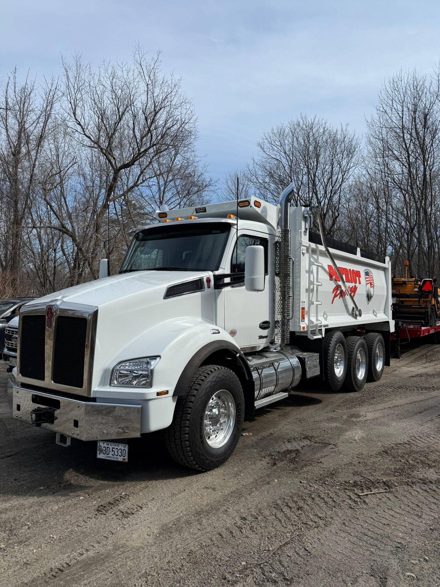 A white Kenworth dump truck parked on a dirt lot, featuring a large chrome bumper and an attached red trailer.