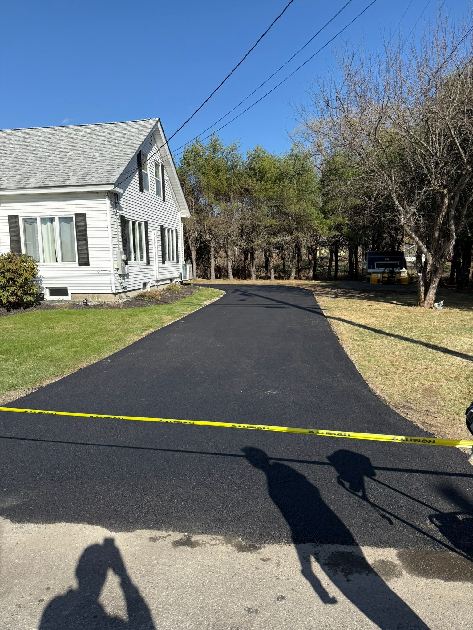 A newly paved black asphalt driveway leads to a white house with a yellow caution tape stretched across the entrance.