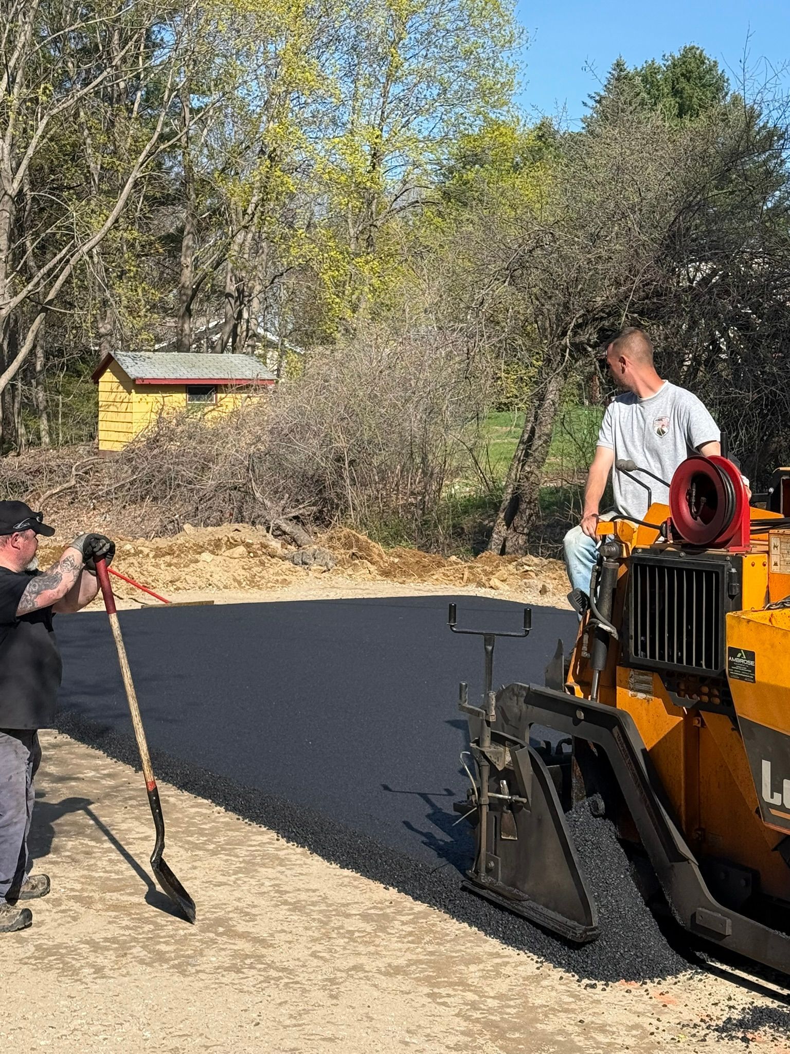 Two people work on a sunny day using a paving machine to lay fresh asphalt on a gravel driveway.
