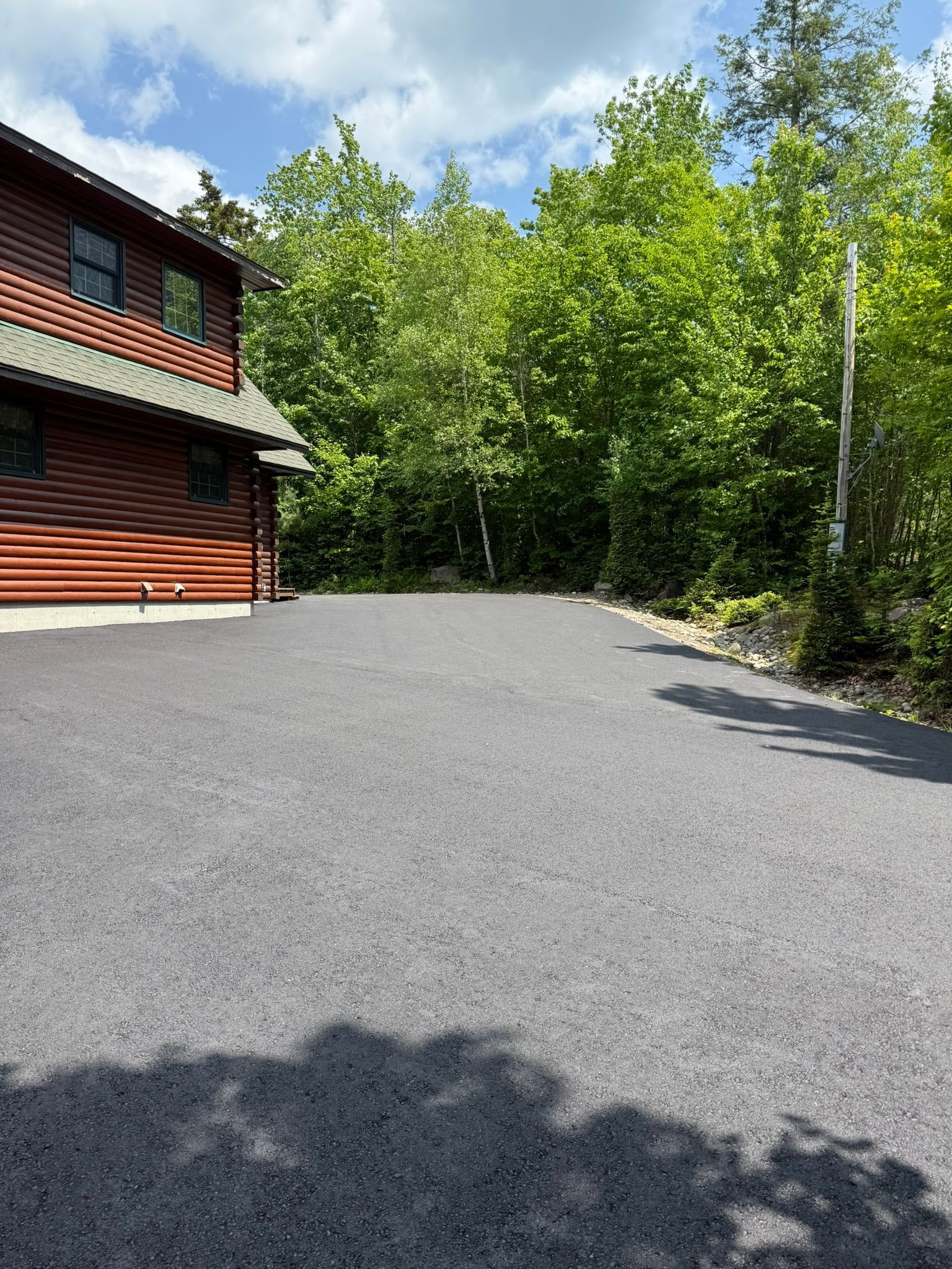 A log cabin with a dark metal roof sits beside a wide, freshly paved gravel driveway, framed by dense green trees.