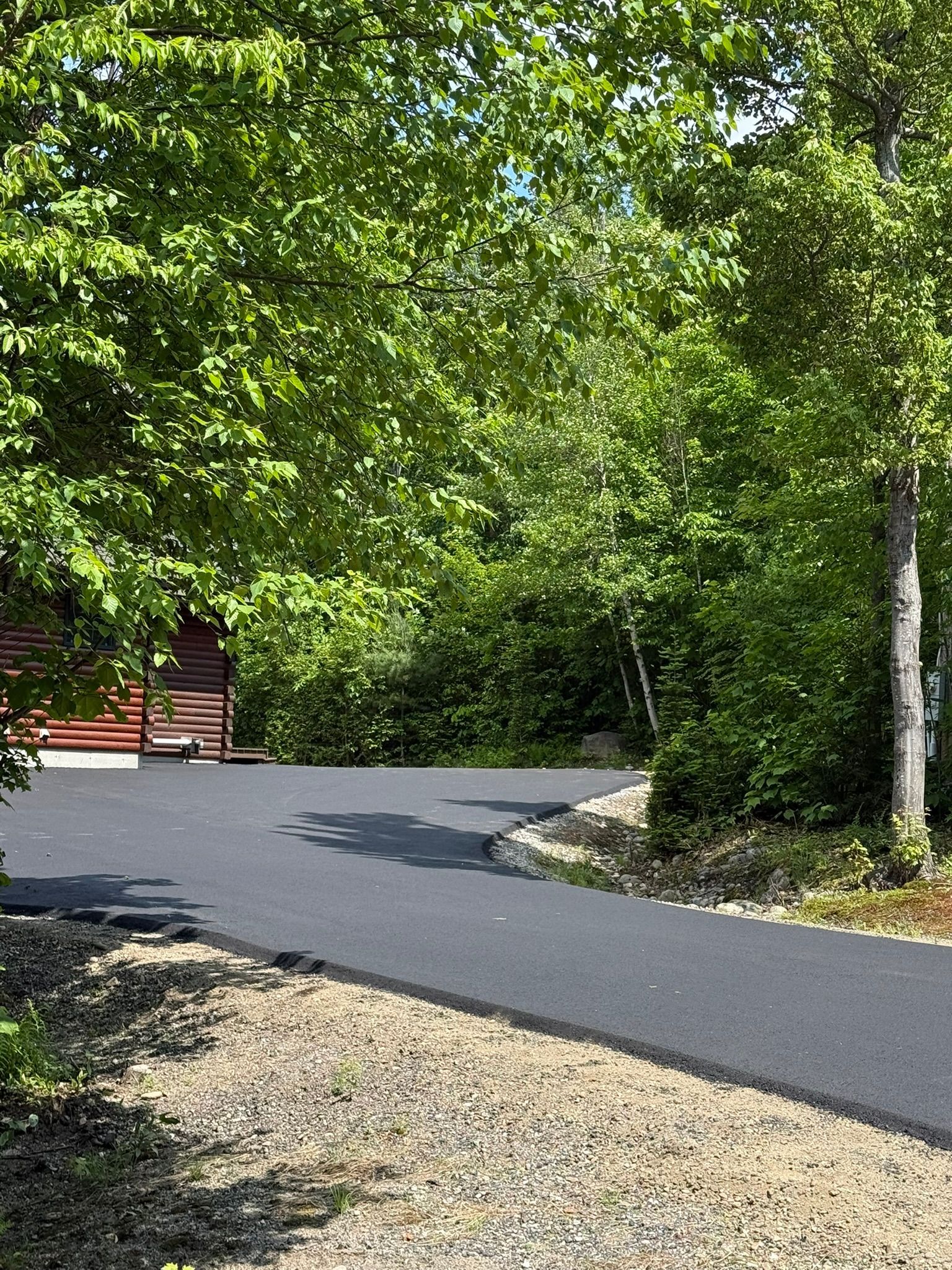 A freshly paved black asphalt driveway curves toward a red wooden building framed by lush green trees.