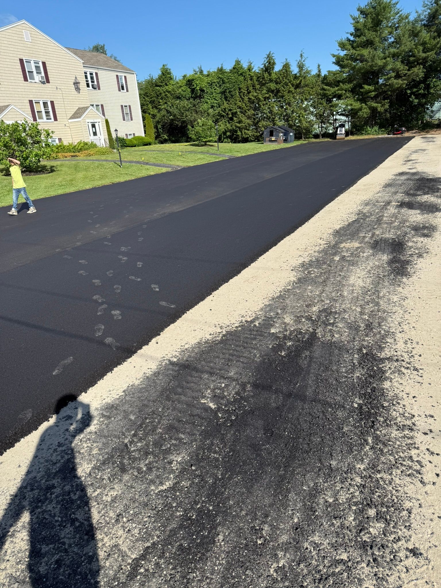 A freshly paved black asphalt driveway adjacent to a strip of loose gravel and dirt near a house on a sunny day.