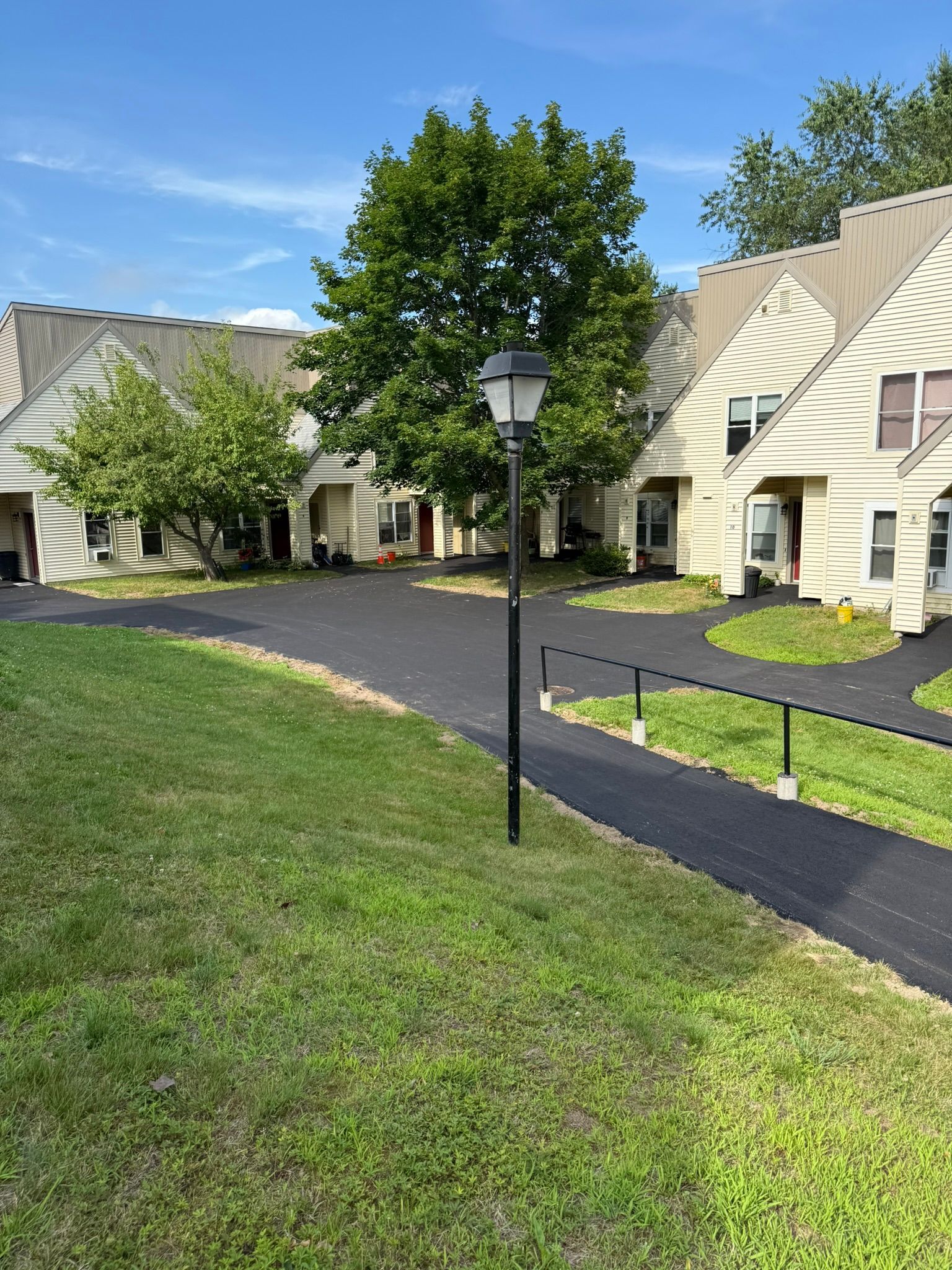 A black metal lamp post stands on a lawn next to a freshly paved black asphalt walkway and driveway near tan townhomes.