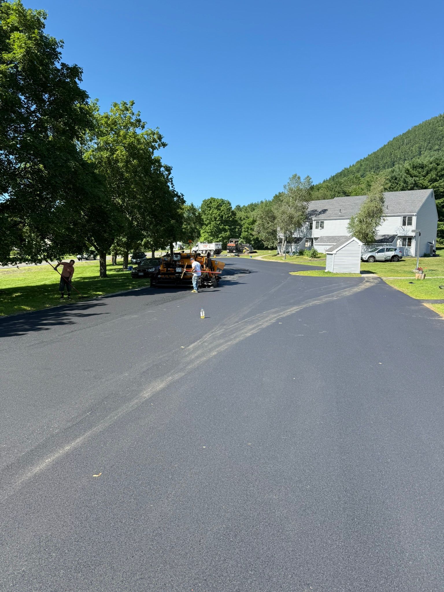 Freshly paved black asphalt driveway leading to a multi-story house near a wooded hill on a sunny day.