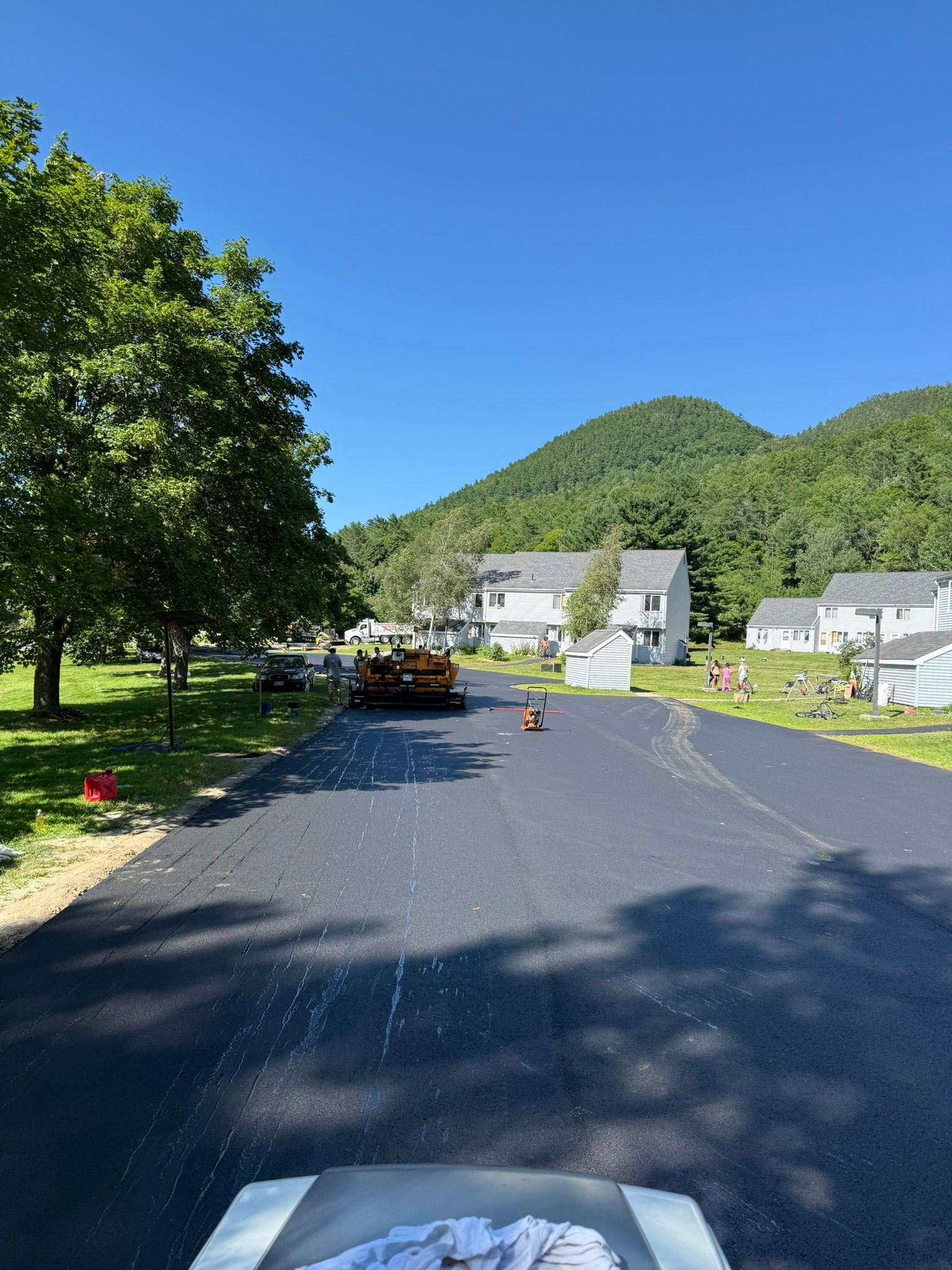 A freshly paved black asphalt road leads toward residential houses with a green mountain in the background.
