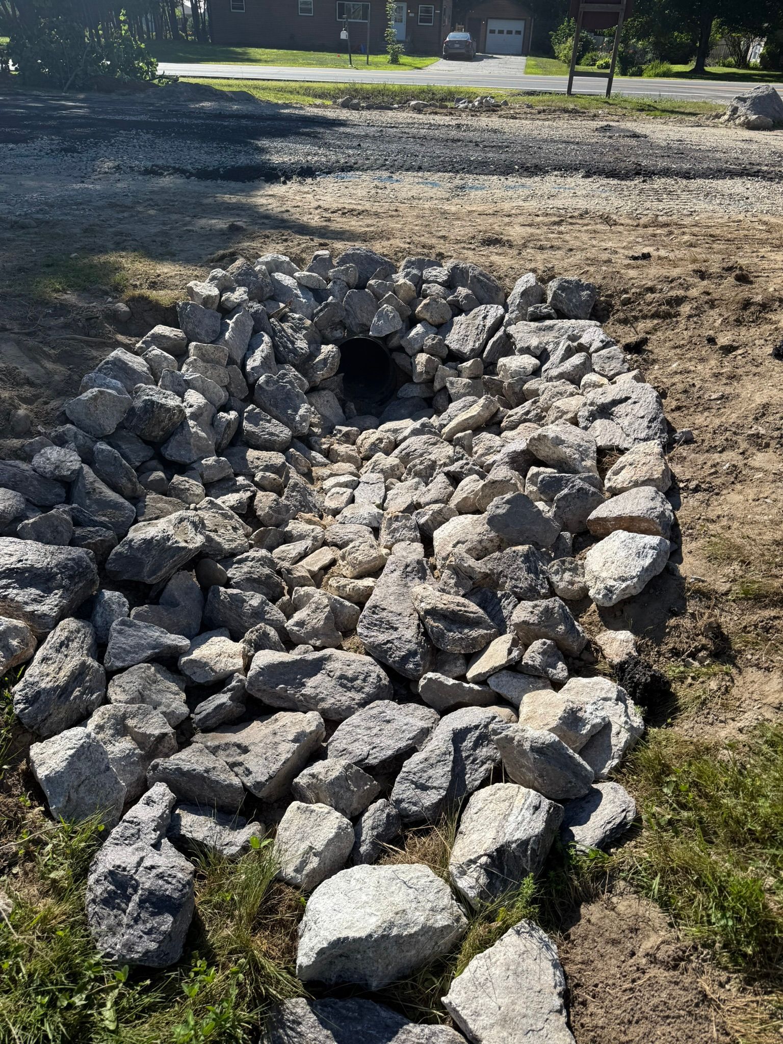 A drainage culvert surrounded by grey riprap stones in a dirt patch near a driveway and house.