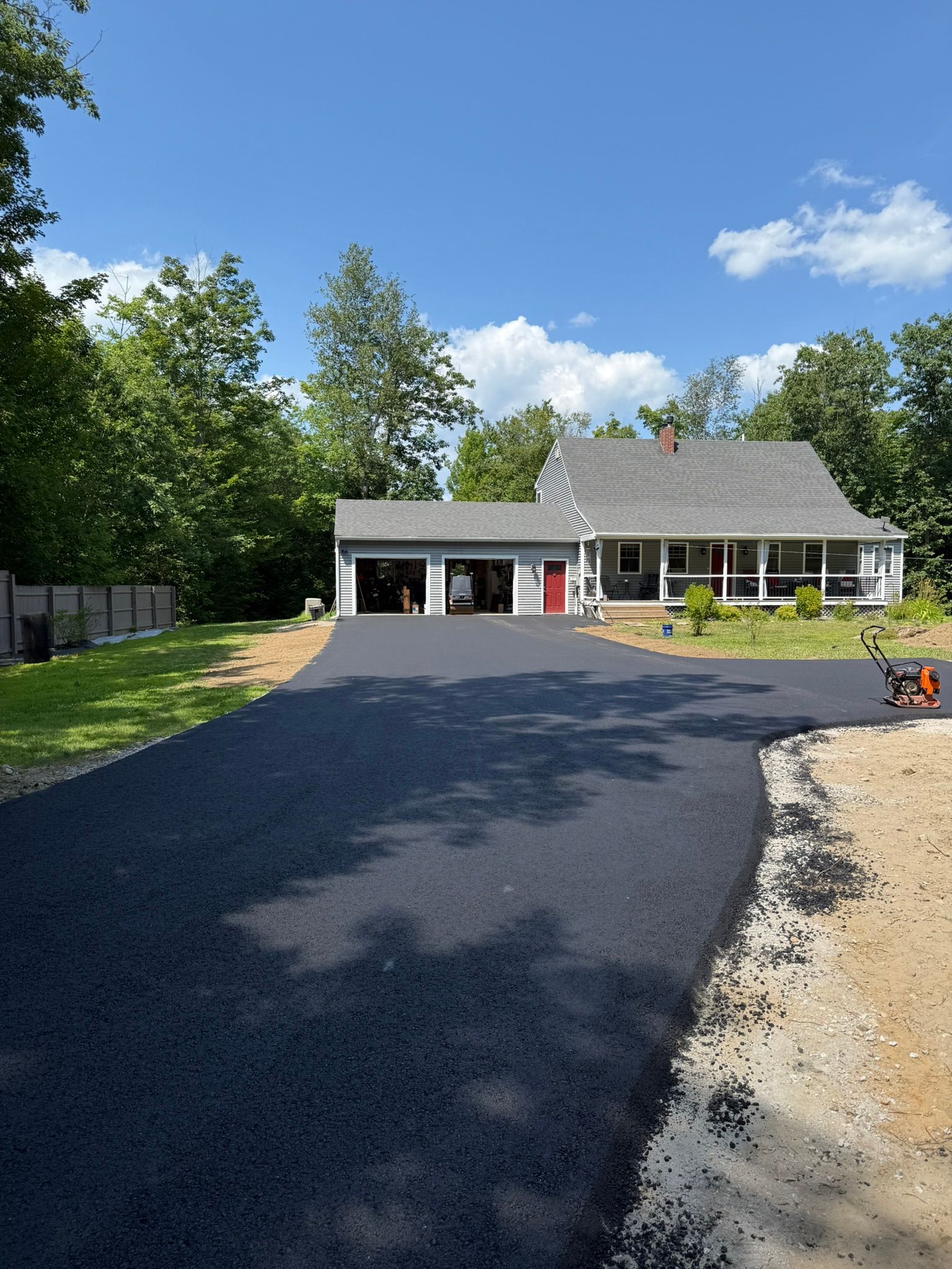 Newly paved asphalt driveway leading to a two-story house with a white porch and attached garage, set among green trees.