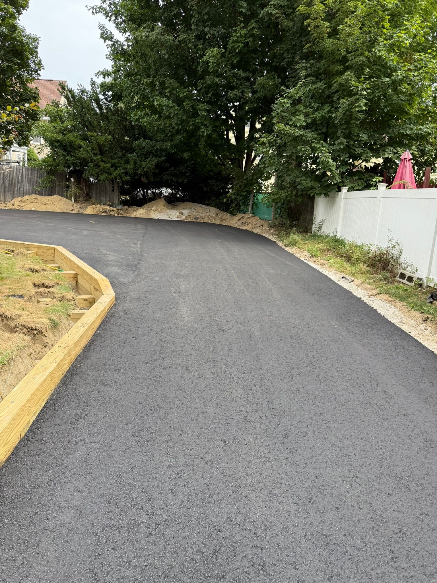 A freshly paved asphalt driveway curves past a wooden raised garden bed toward trees and a white fence.