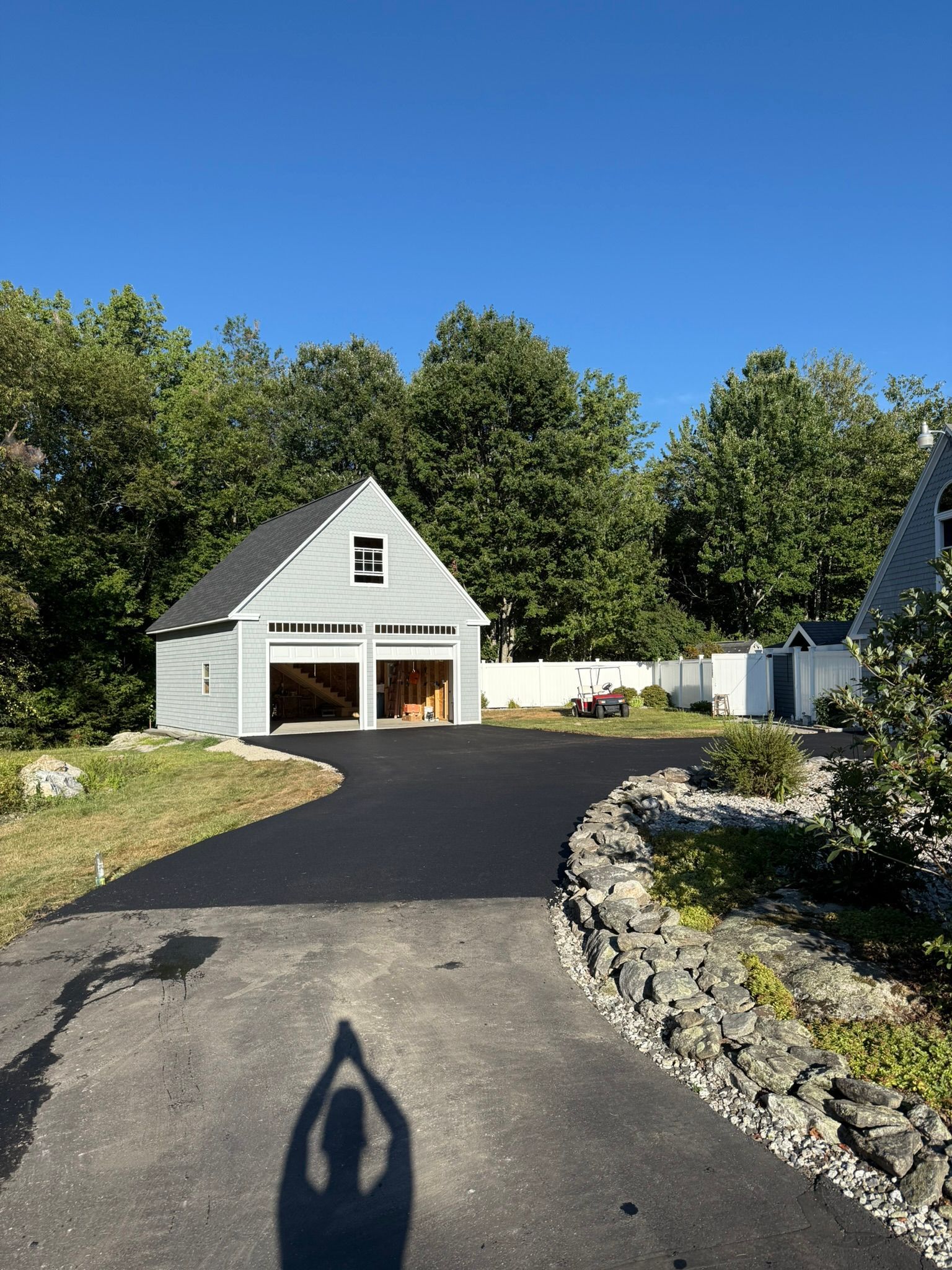 A light grey garage with two open bays at the end of a freshly paved asphalt driveway, surrounded by trees and a stone wall.