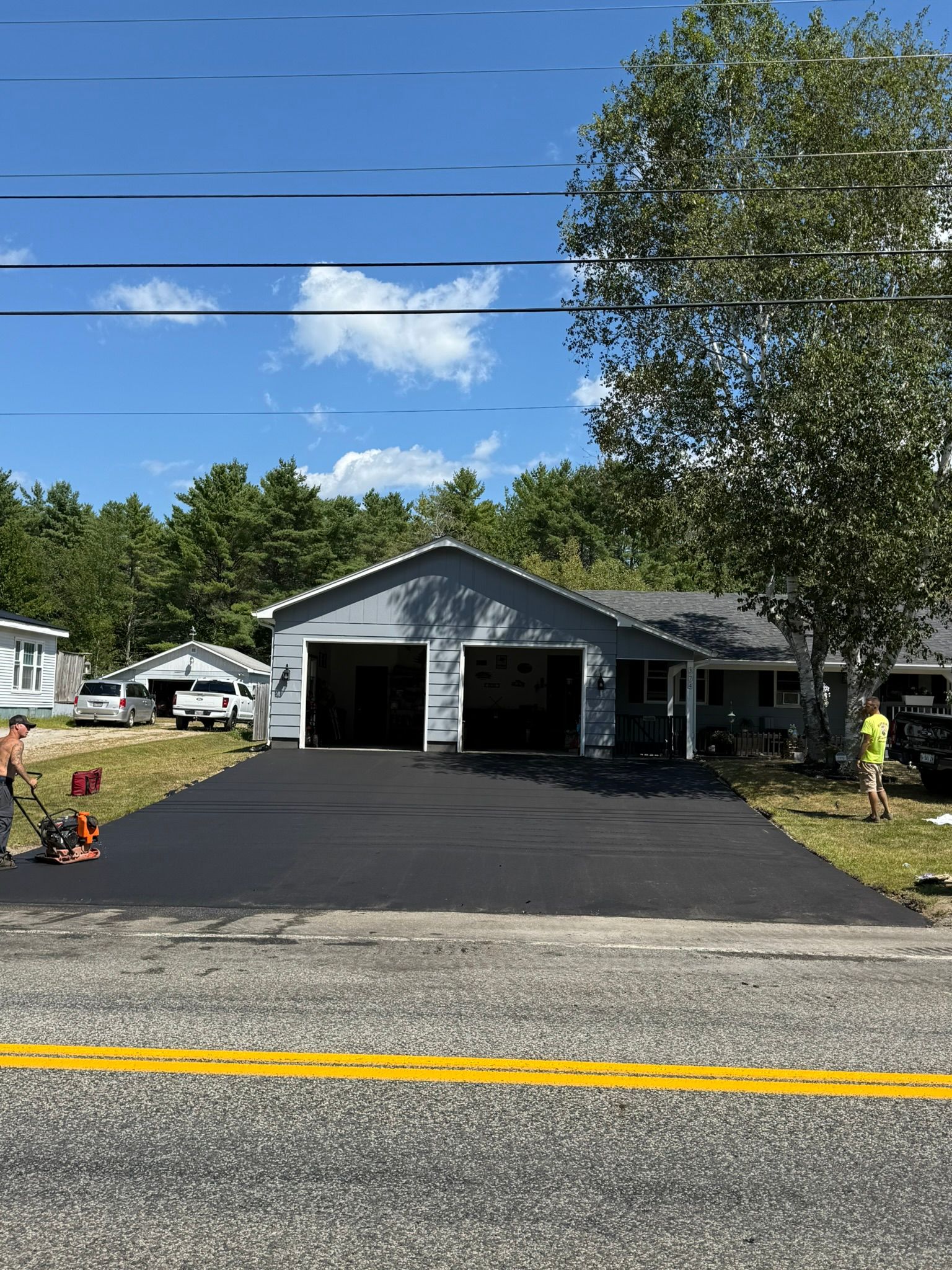 Workers pave a fresh black asphalt driveway in front of a gray residential house on a sunny day.
