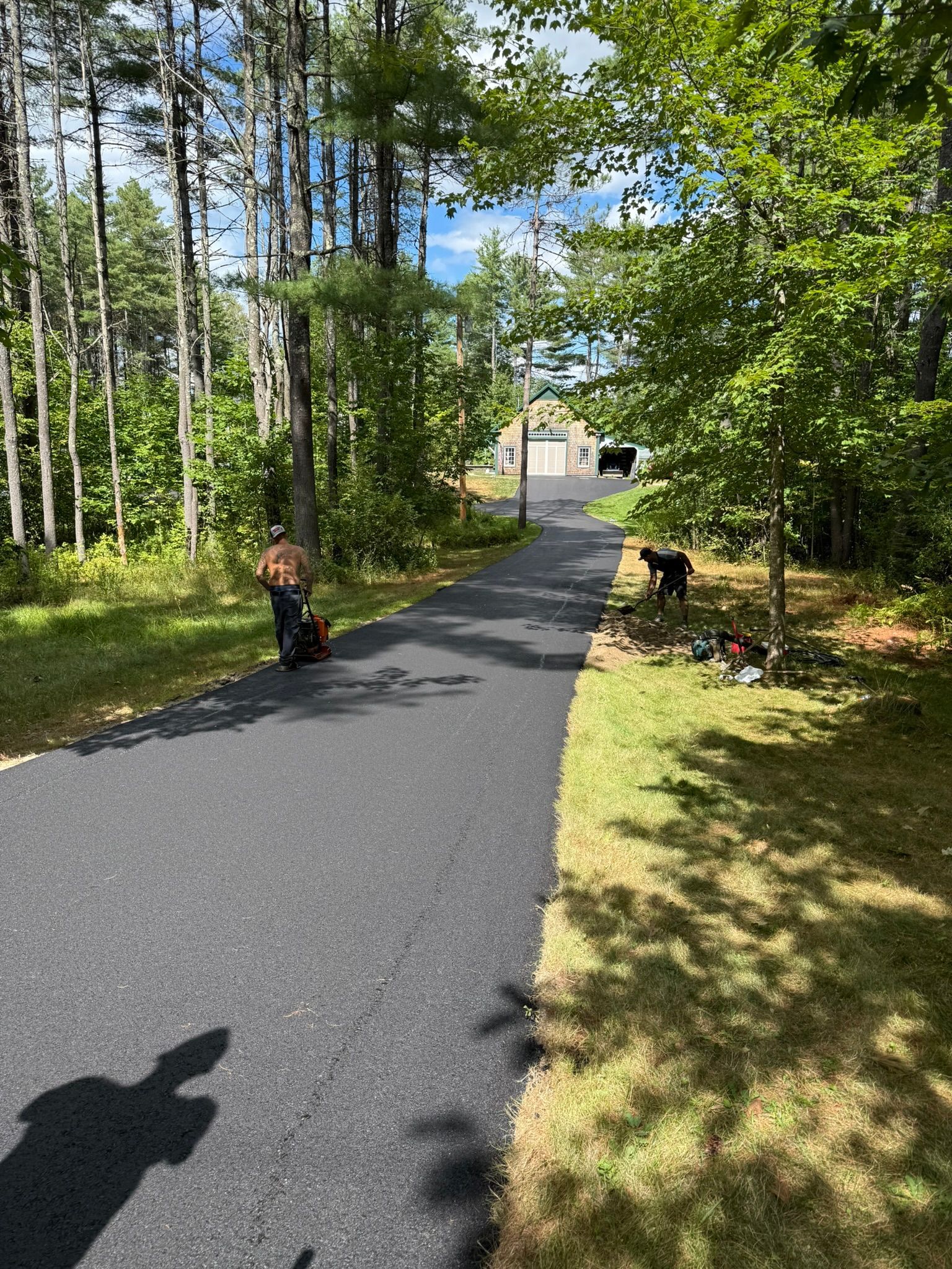 A freshly paved, dark asphalt driveway leads through a wooded, sunny landscape toward a house in the distance.