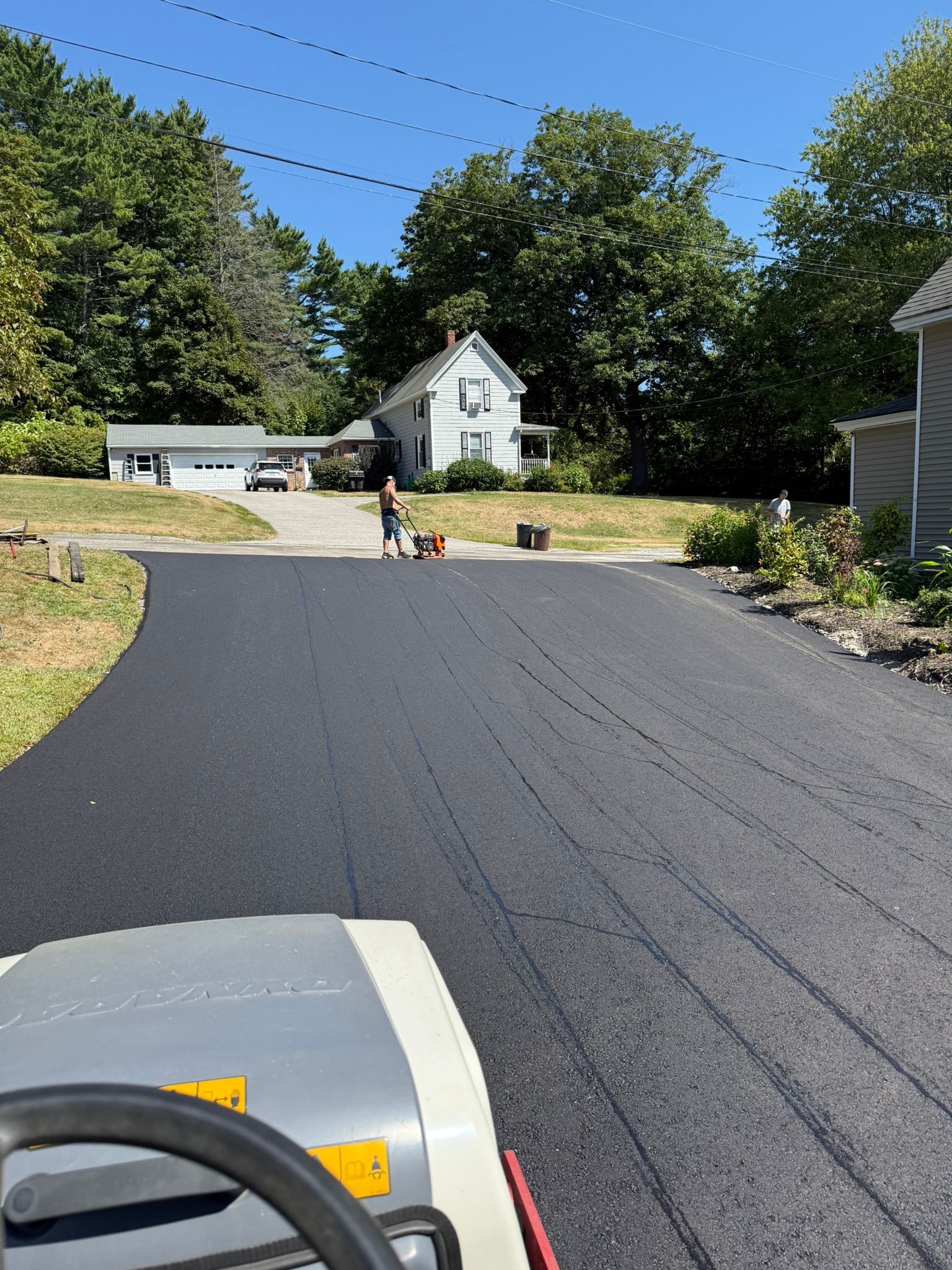 A view from a construction roller onto a freshly paved asphalt driveway, with a person operating equipment in the distance.