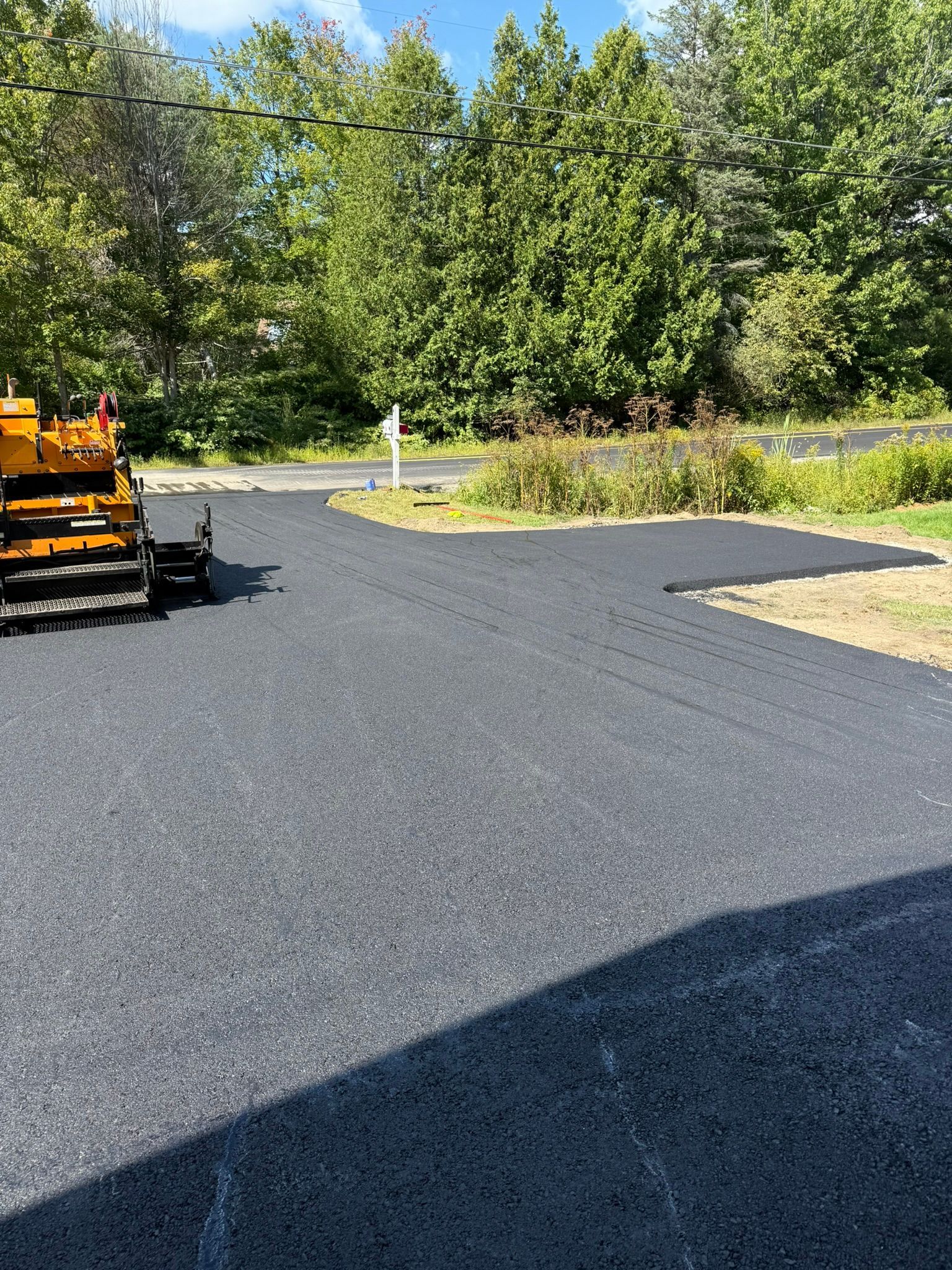 A yellow construction vehicle sits at the edge of a freshly paved, smooth black asphalt driveway lined by green trees.