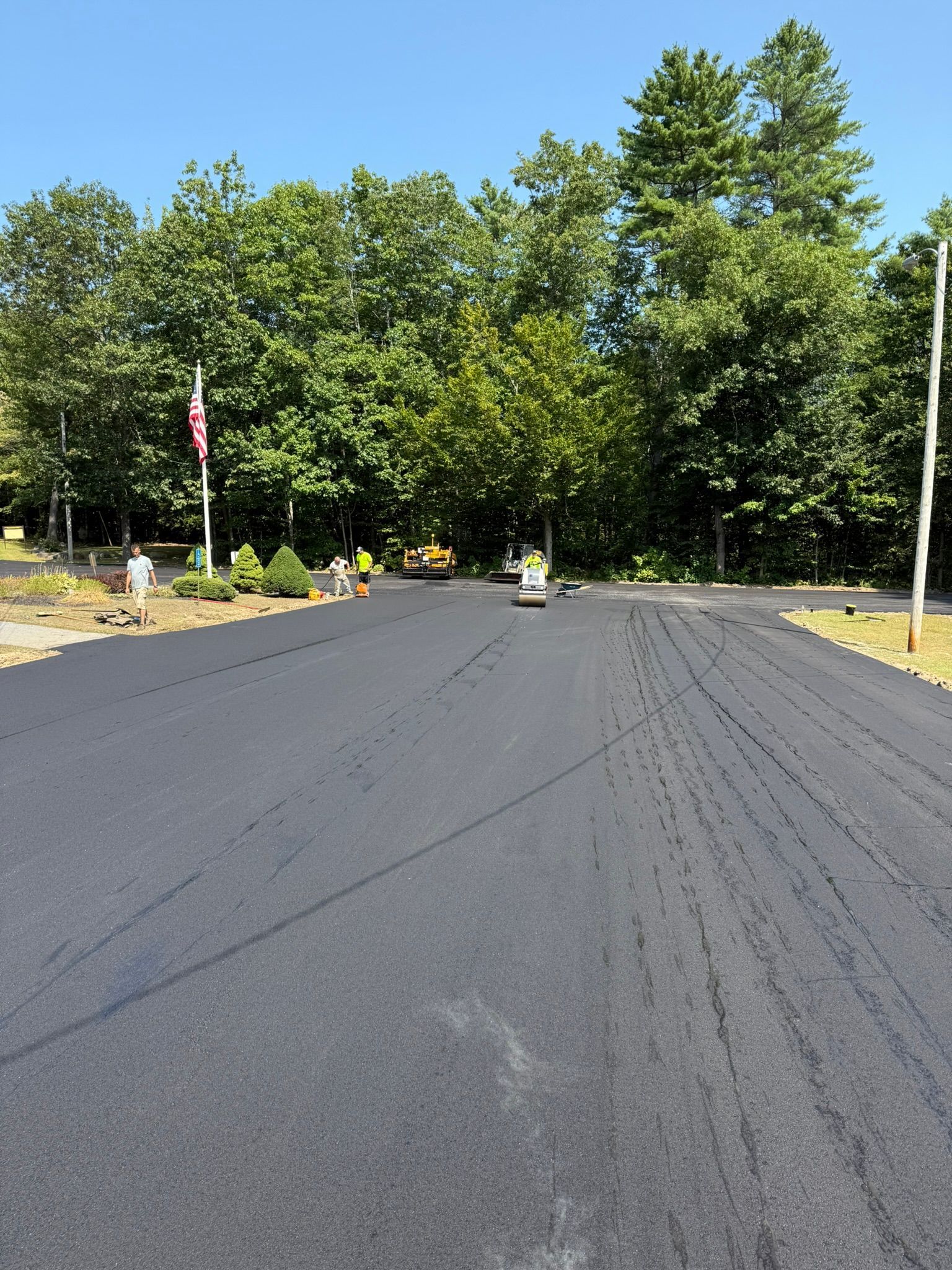 Workers pave a fresh asphalt driveway leading to a tree-lined lot on a sunny day.