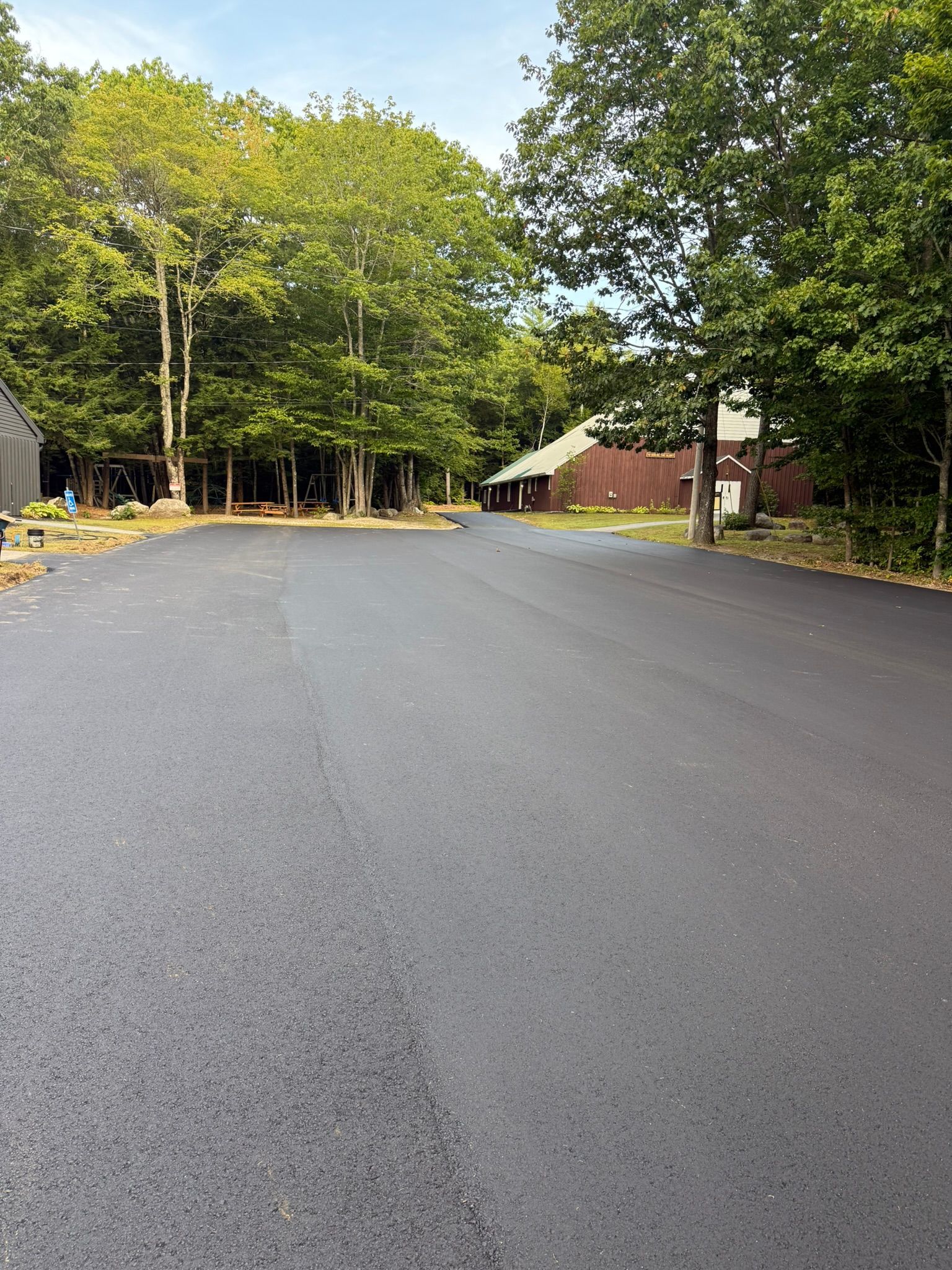 A wide, freshly paved black asphalt road leads toward a brown building set amongst a backdrop of lush green trees.