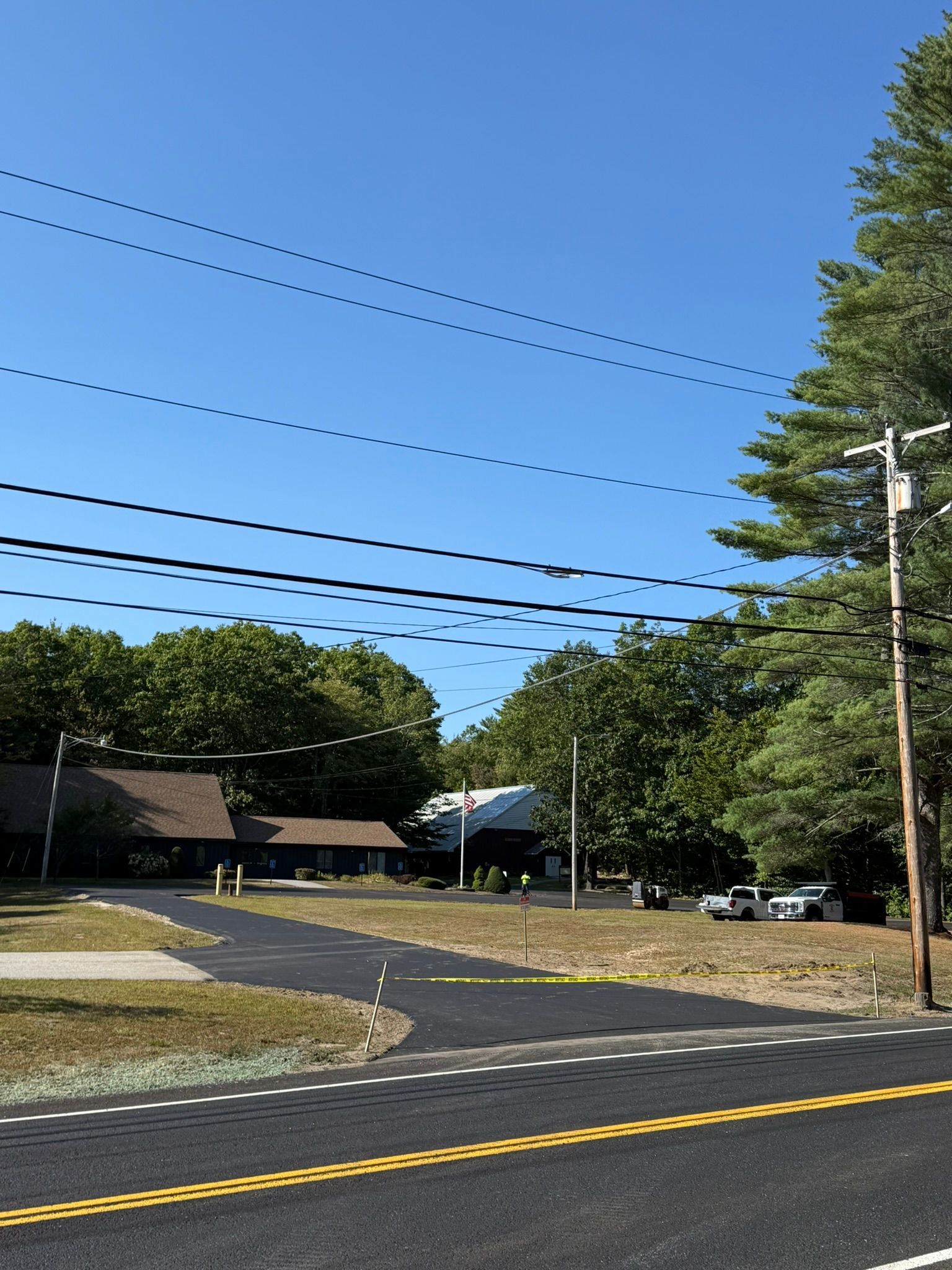 A wide, freshly paved asphalt parking lot leads toward a brown, A-frame building surrounded by dense green trees.